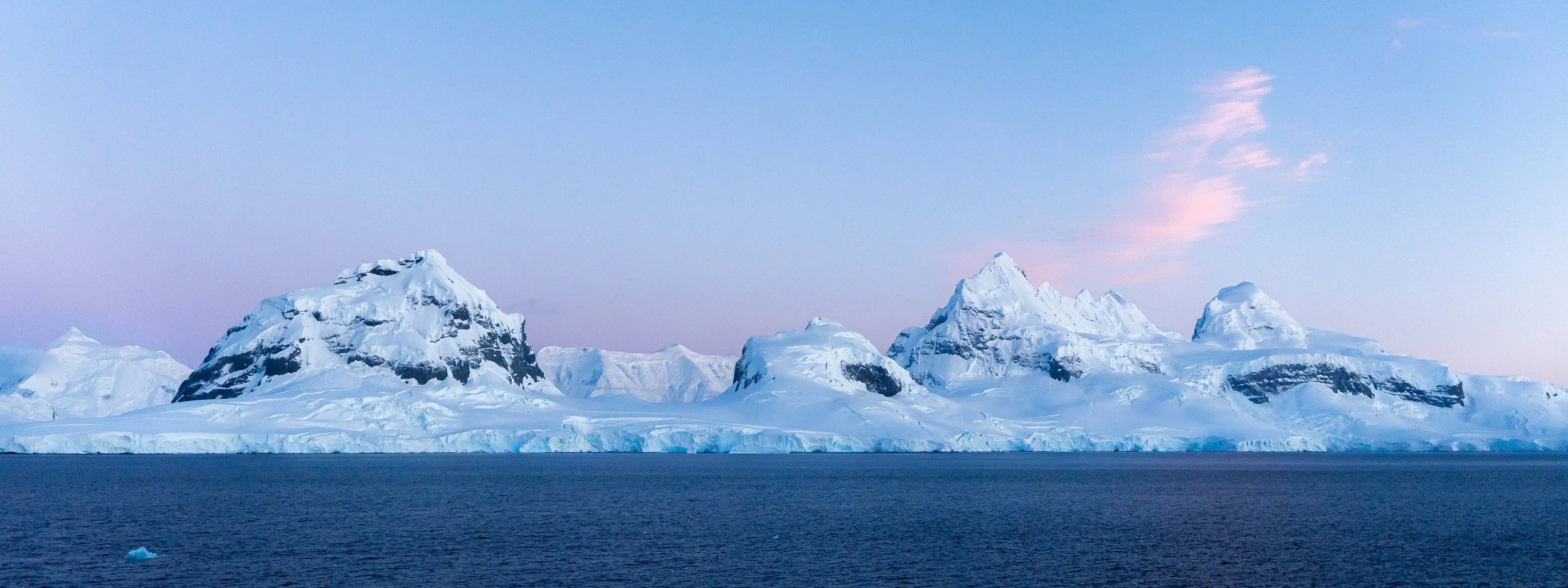 A group of snow-covered mountains and icebergs in the ocean under a pastel-colored sky.