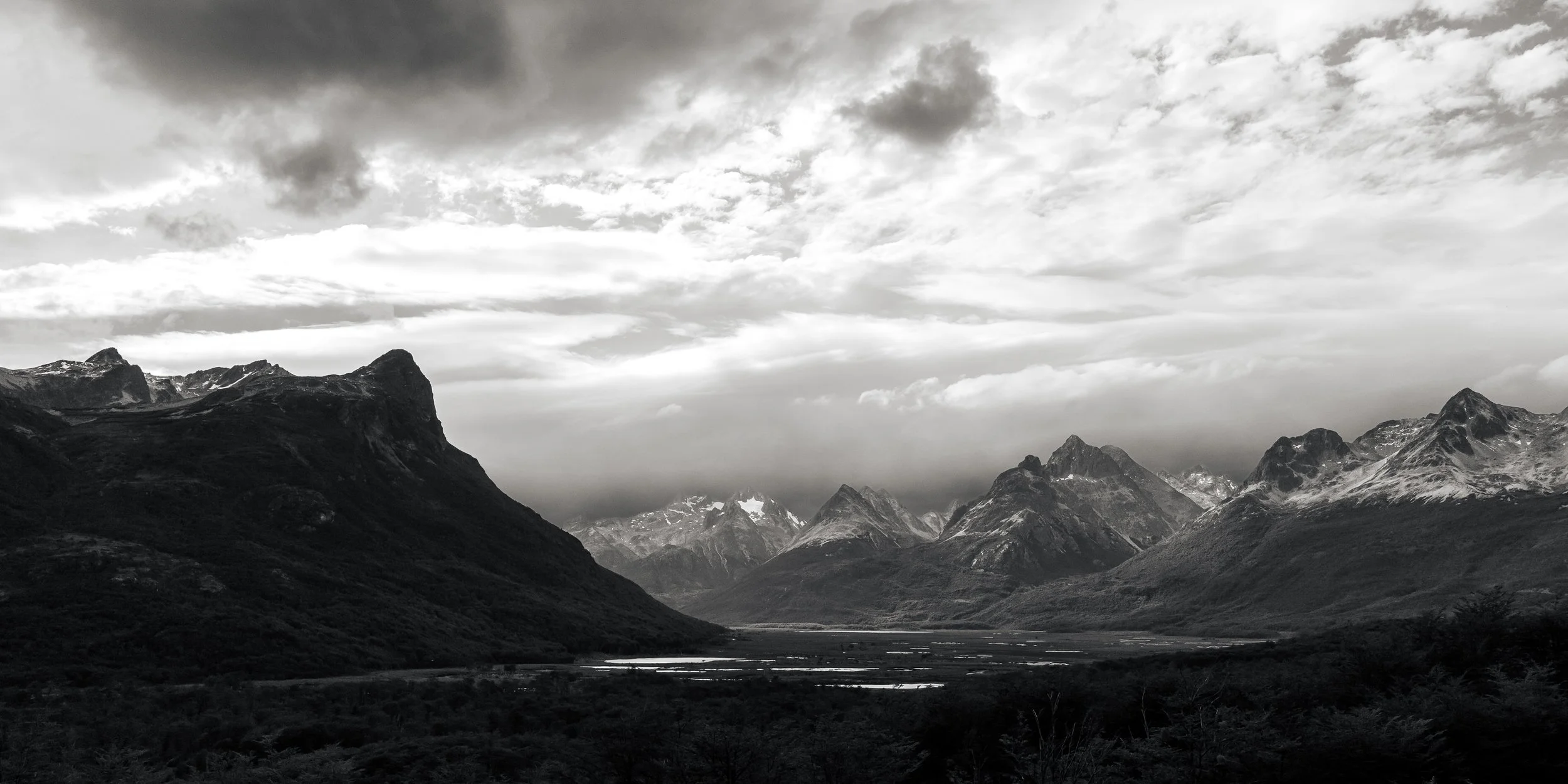 A black and white photo of a mountainous landscape with rugged peaks, a cloudy sky, and a valley with water bodies in the foreground.