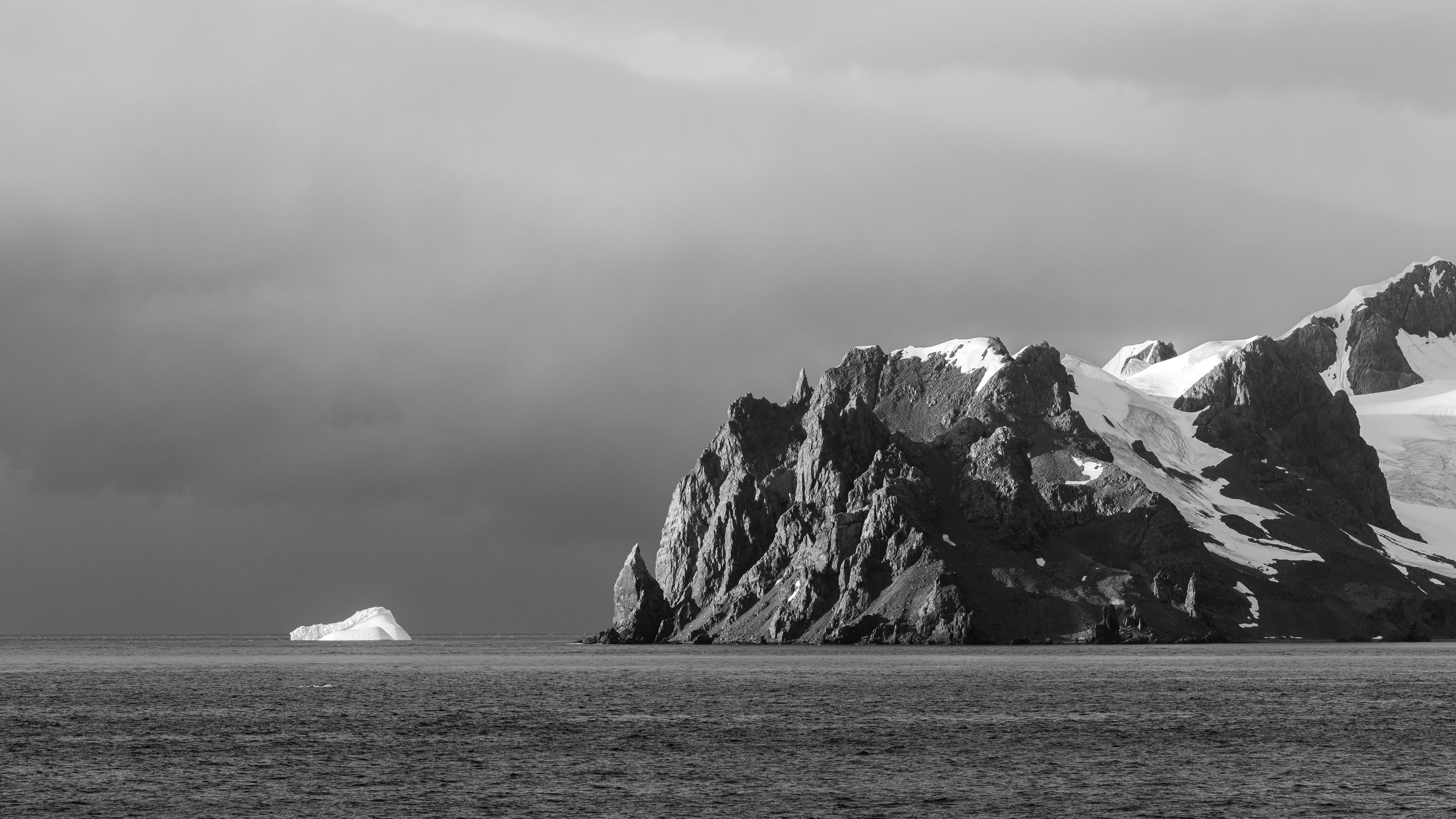 Black and white photograph of rugged mountainous coastline with snow-capped peaks, a large rock formation in the sea, and an iceberg near the coast under a cloudy sky.