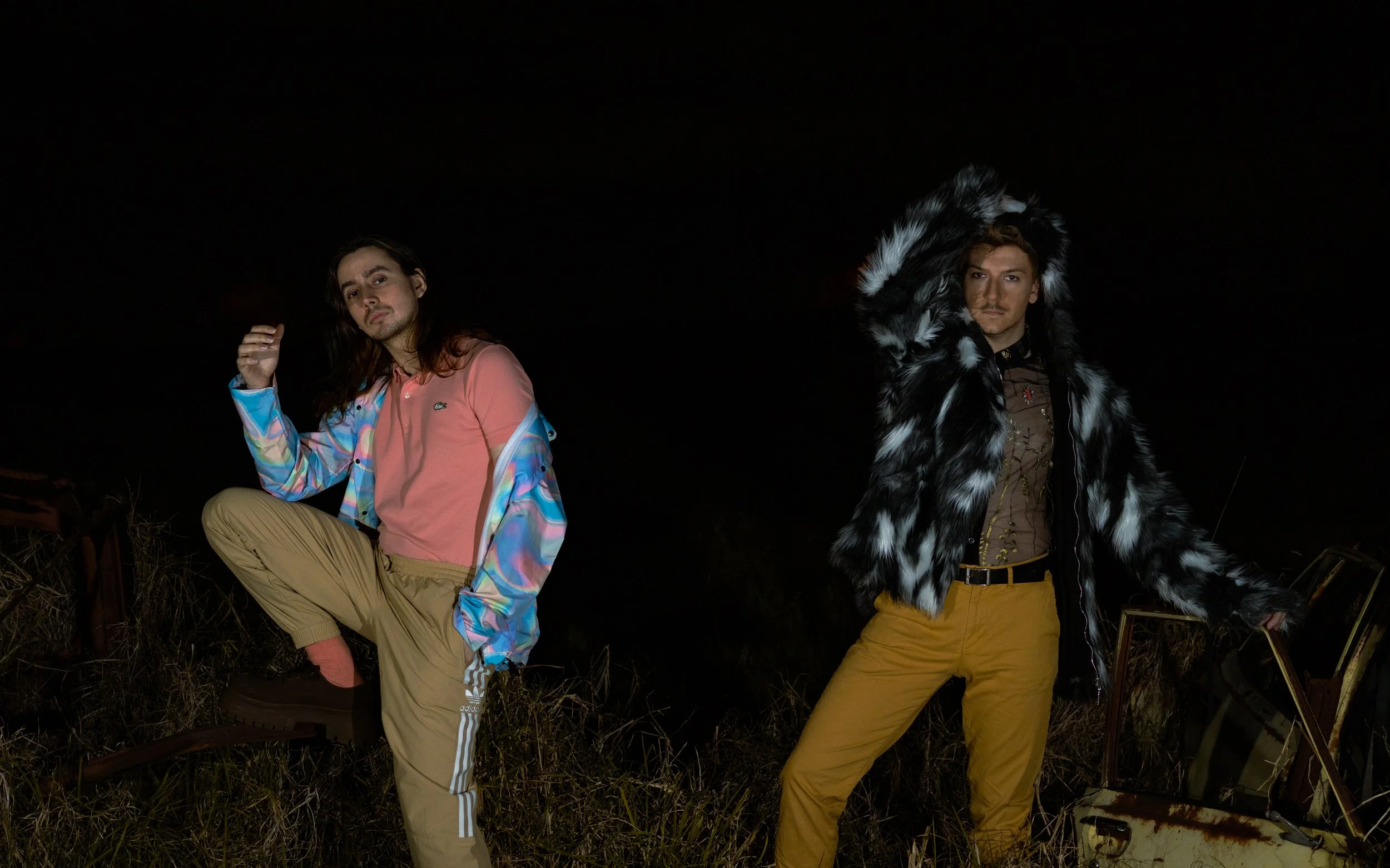 Two young men posing outdoors at night with a dark background. One is kneeling with long hair, wearing a colorful windbreaker and beige pants, while the other is standing with short hair, wearing a fur coat and mustard-colored pants.