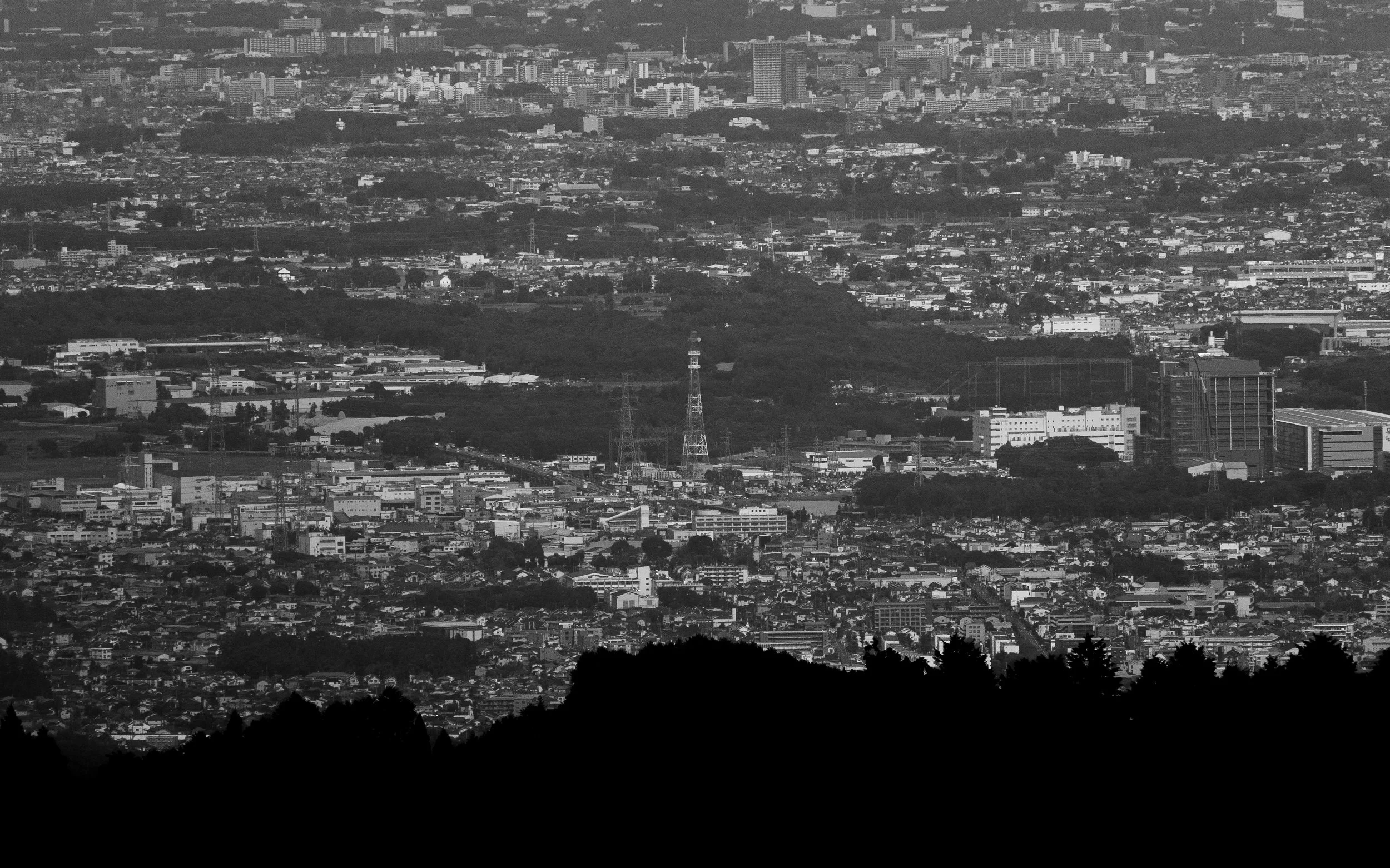 A black-and-white aerial view of a large city with numerous buildings, high-rises, and a network of roads. The landscape includes a mix of densely packed urban areas and some green spaces or parks. Notable structures include transmission towers visib