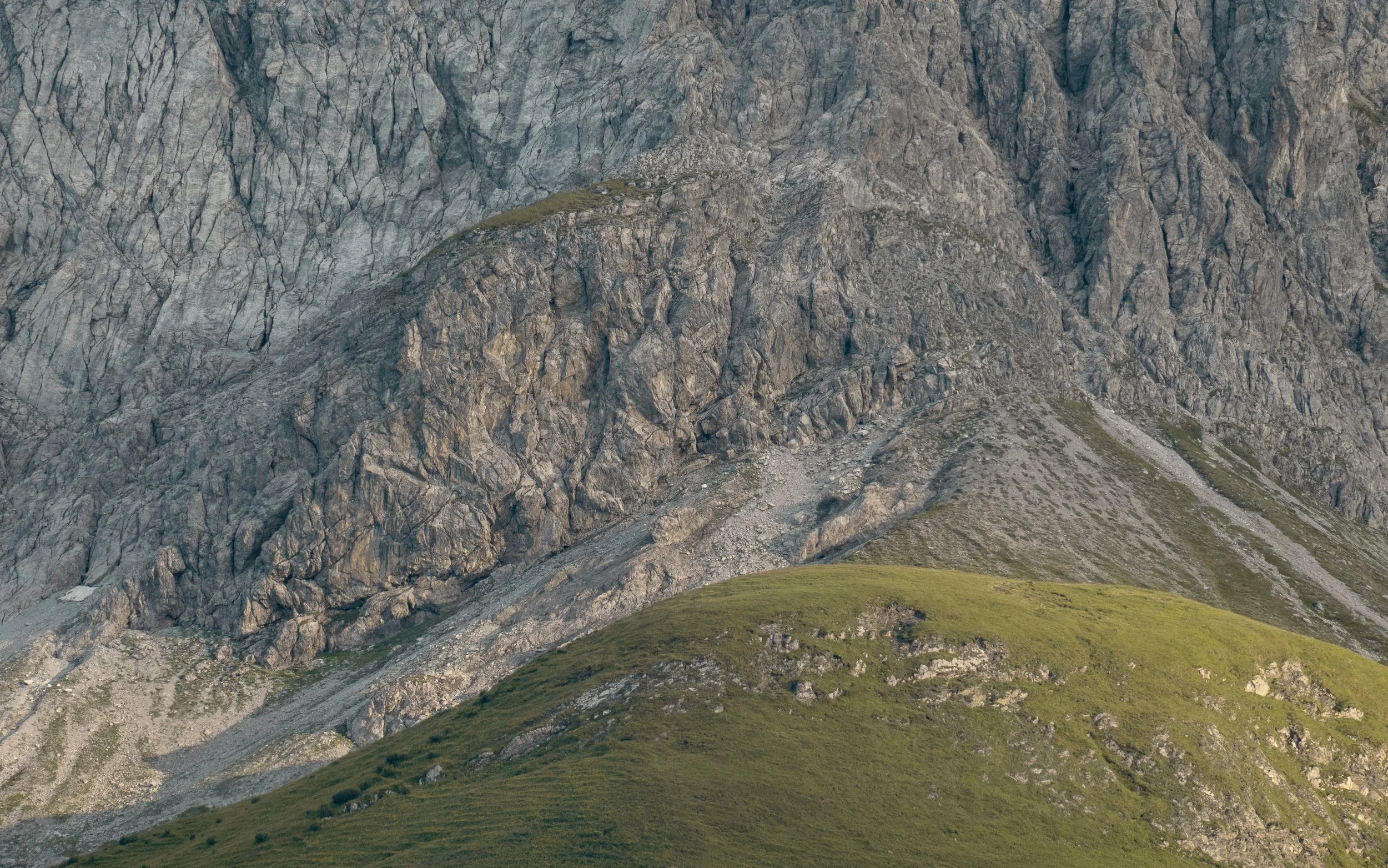 A rugged mountain face with steep, rocky cliffs and patches of green grass at the base.