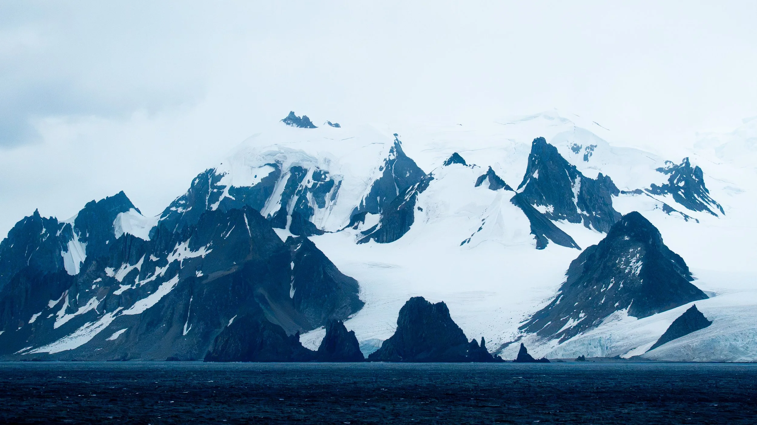 Snow-covered mountain range with dark rocky peaks and a glacier, under a cloudy sky. Body of water in the foreground.