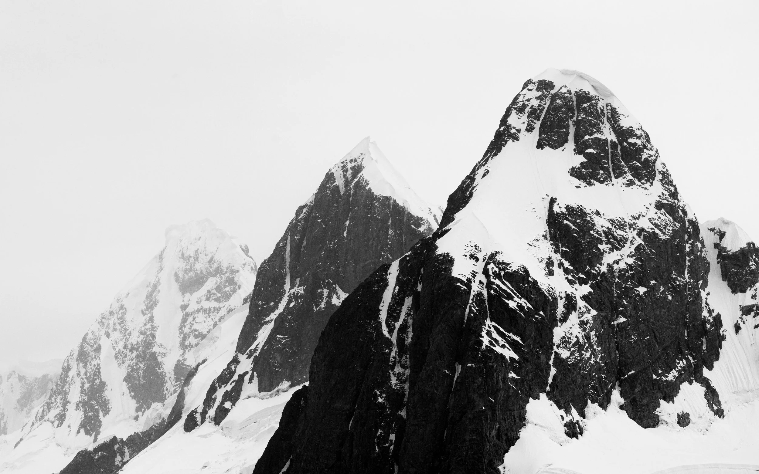 Black and white photo of snow-covered mountain peaks with steep rocky faces, some partially covered in snow, against an overcast sky.