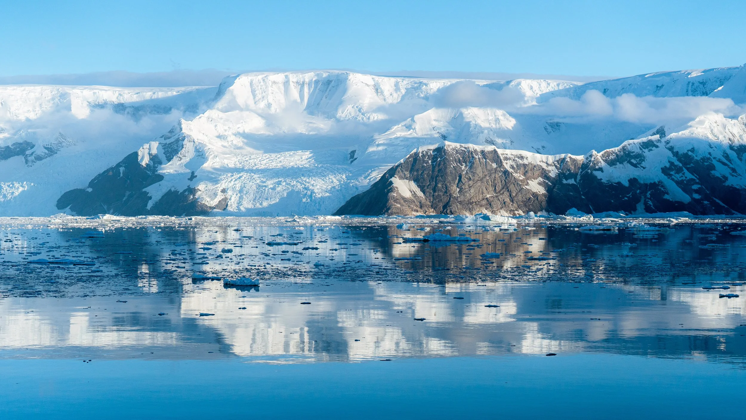 A icy landscape of glaciers and snow-covered mountains reflected in calm water with small ice chunks floating on the surface.