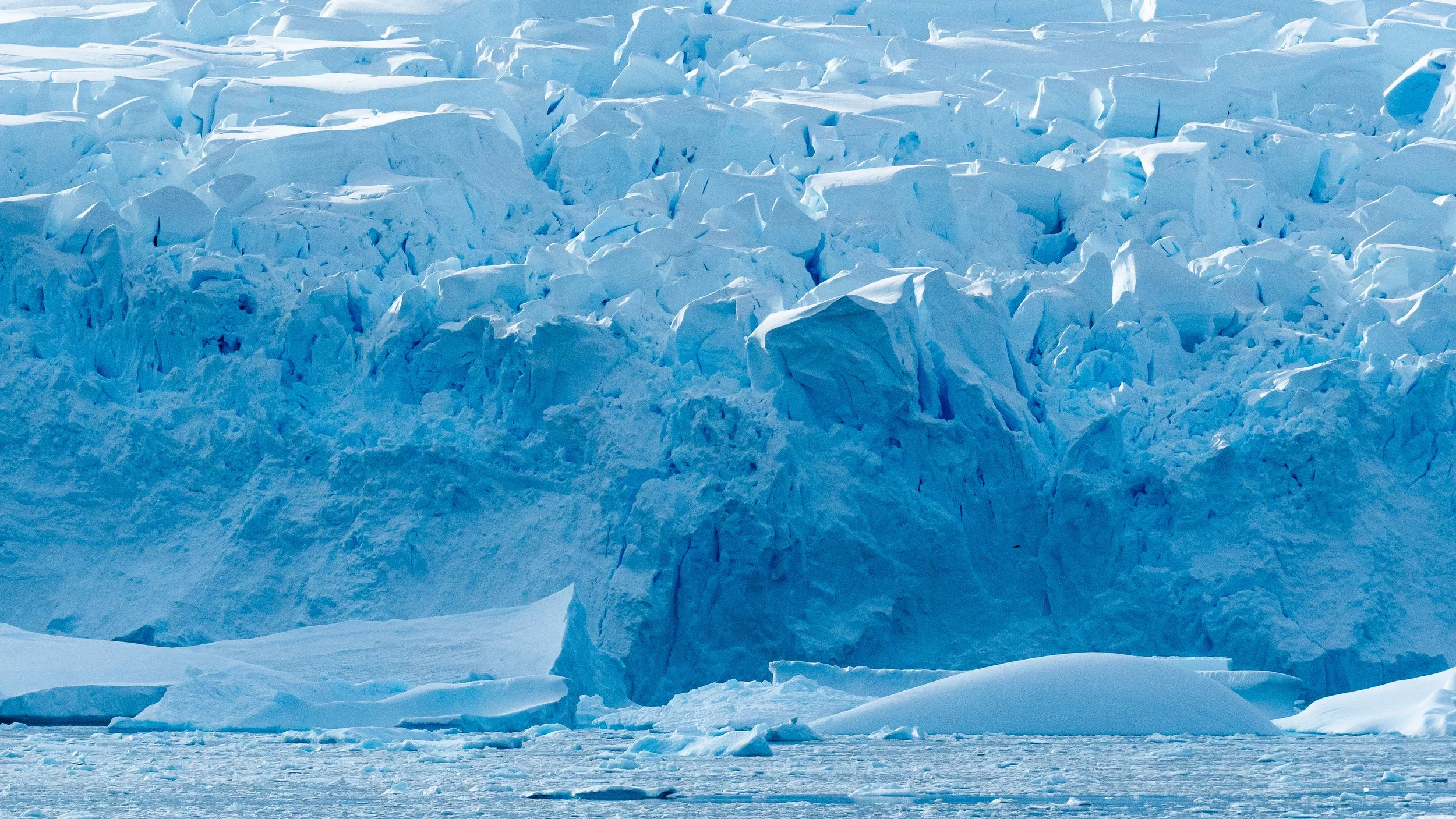 A vast expanse of ice and snow with large broken ice chunks and a glacier in the background under a clear sky.