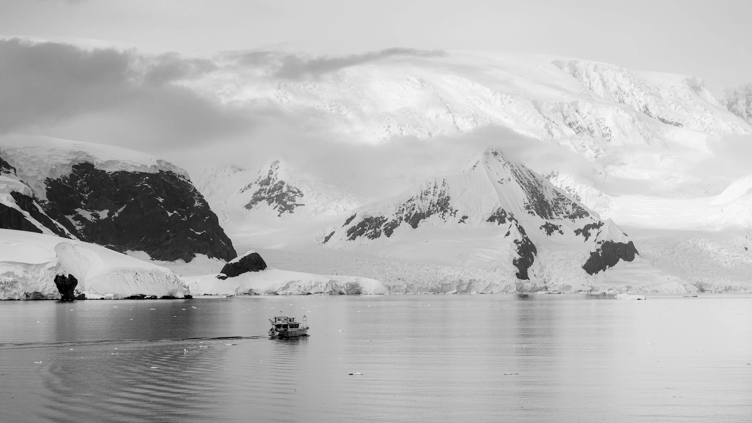 A black and white photo of a boat on calm icy water, with large snow-covered mountains and glaciers in the background.