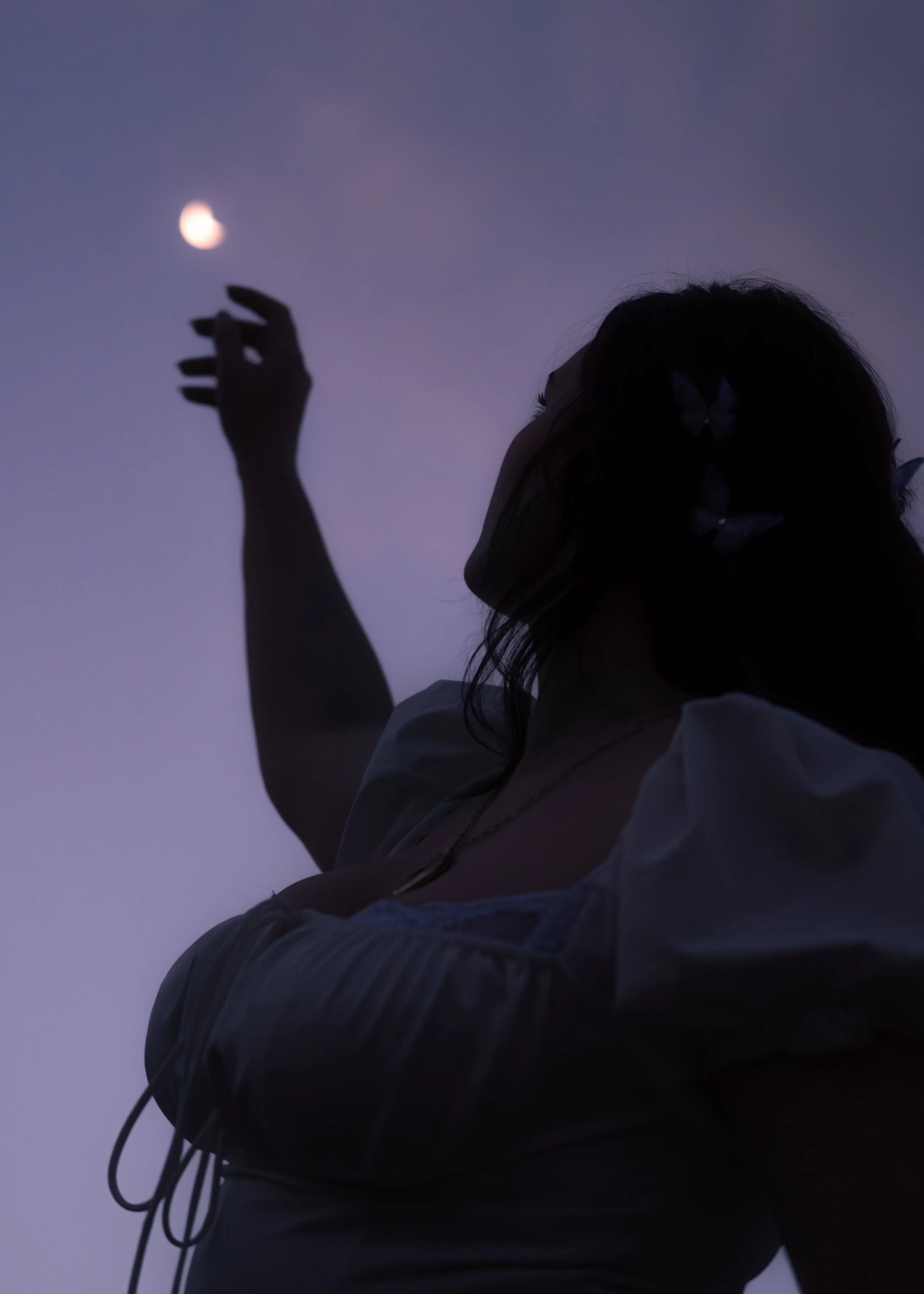 A person with long hair, wearing glasses and a white top, is reaching upward towards a full moon in the evening sky.