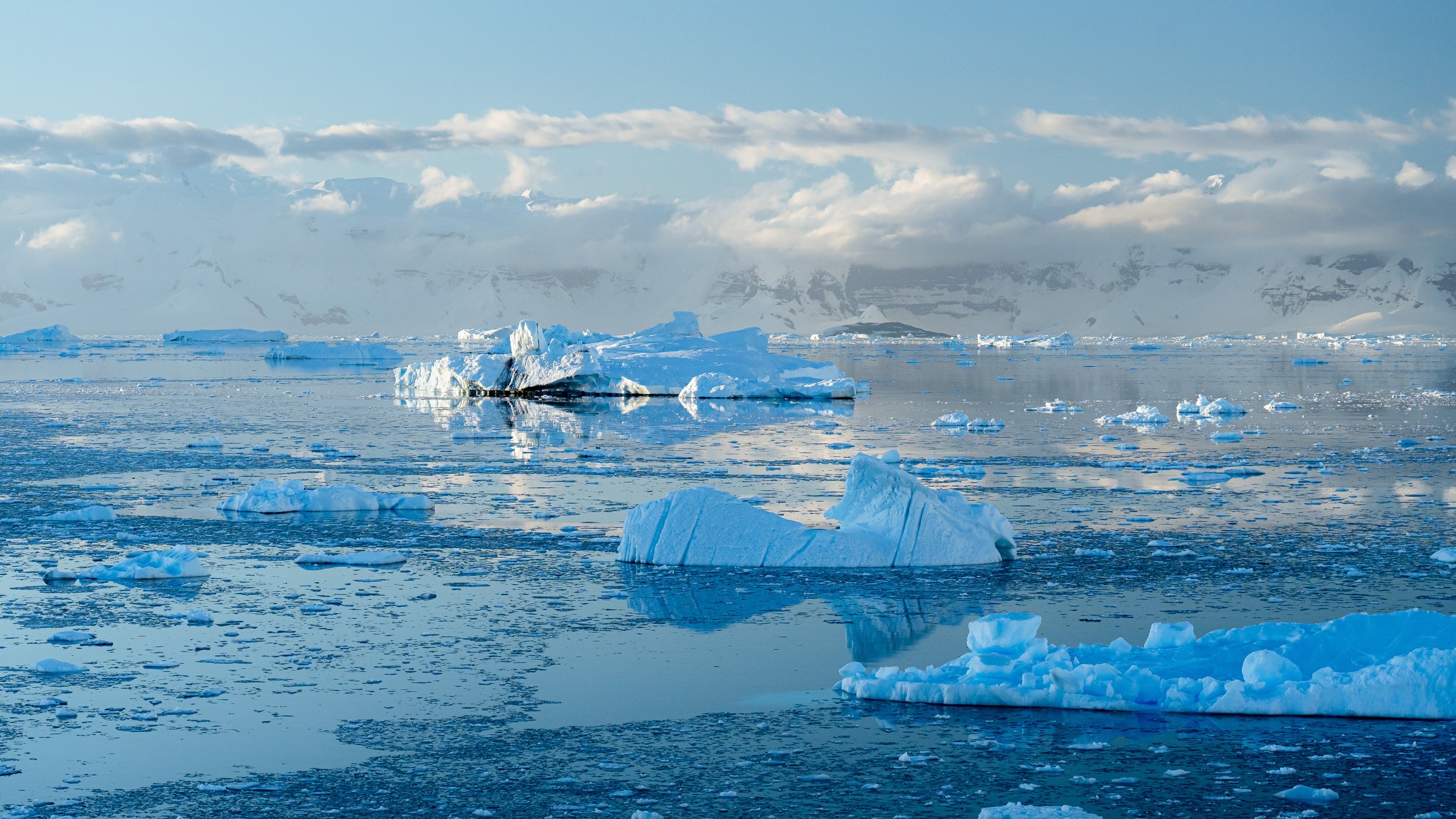 Arctic scene with floating icebergs on calm cold water, snow-covered mountains in the background, and partly cloudy skies.