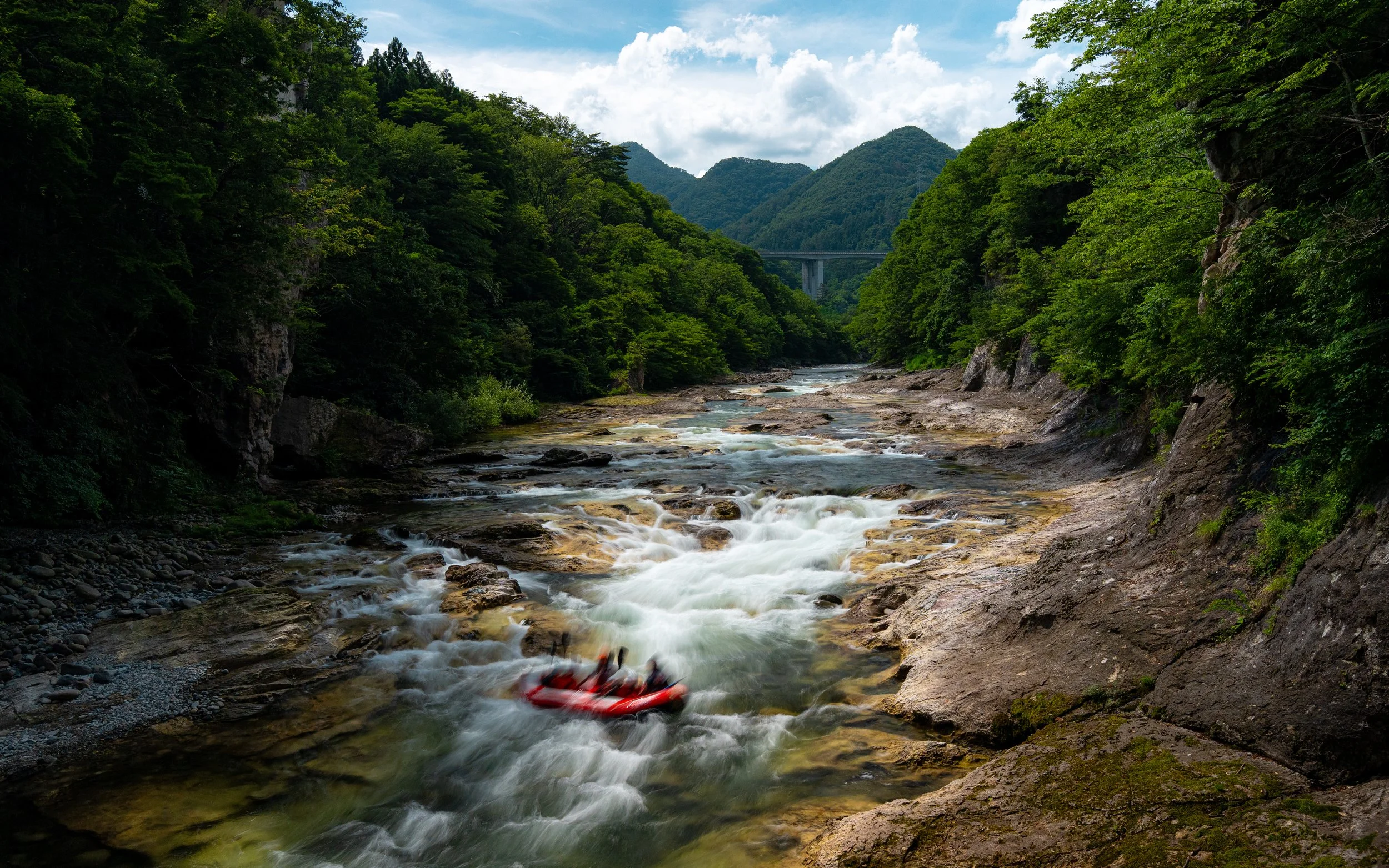 A river flowing through a forested valley with a bridge visible in the background, surrounded by green trees and mountains under a partly cloudy sky.