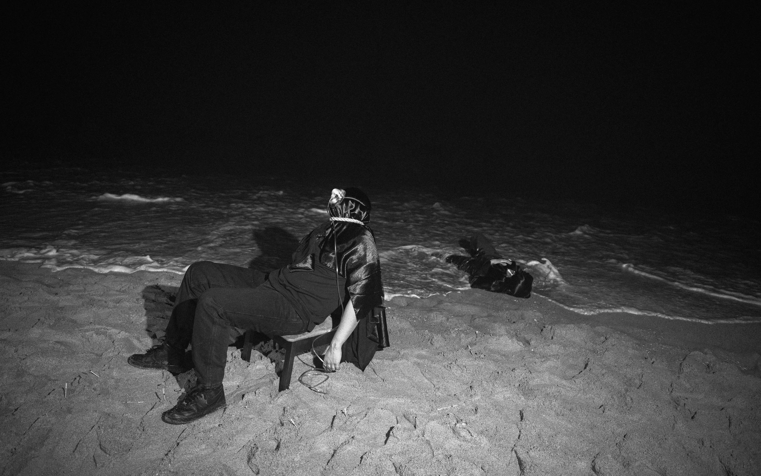 A person sitting on the sand at the beach at night, looking at the water with a dog lying nearby. It is in black and white.