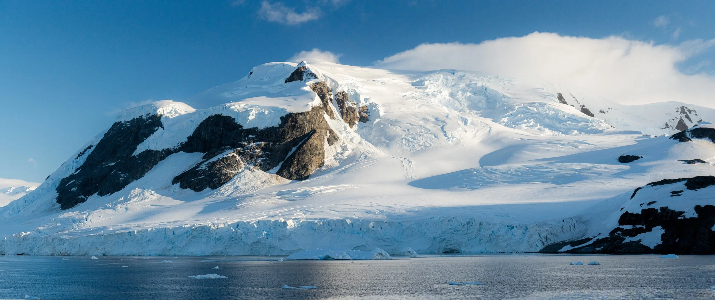 Snow-covered mountain with dark rocky outcrops, ice, and snow, overlooking cold water with floating icebergs under a partly cloudy blue sky.