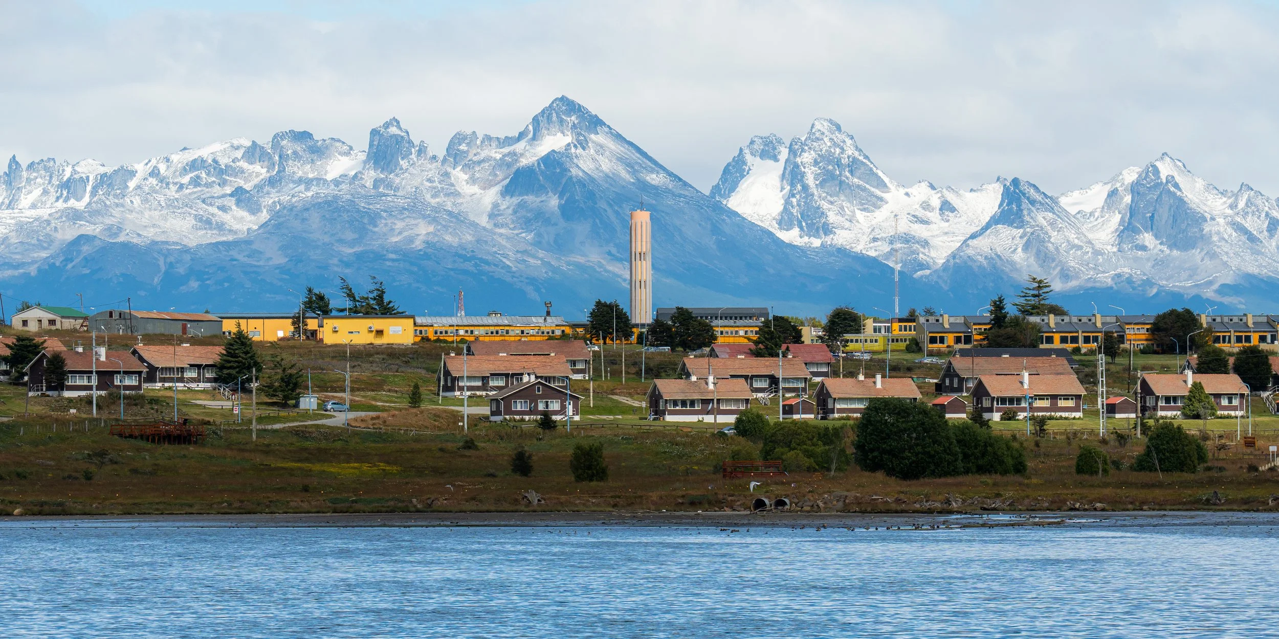 A scenic view of a residential neighborhood with houses, a water tower, and a yellow train in front of a mountain range with snow-capped peaks.