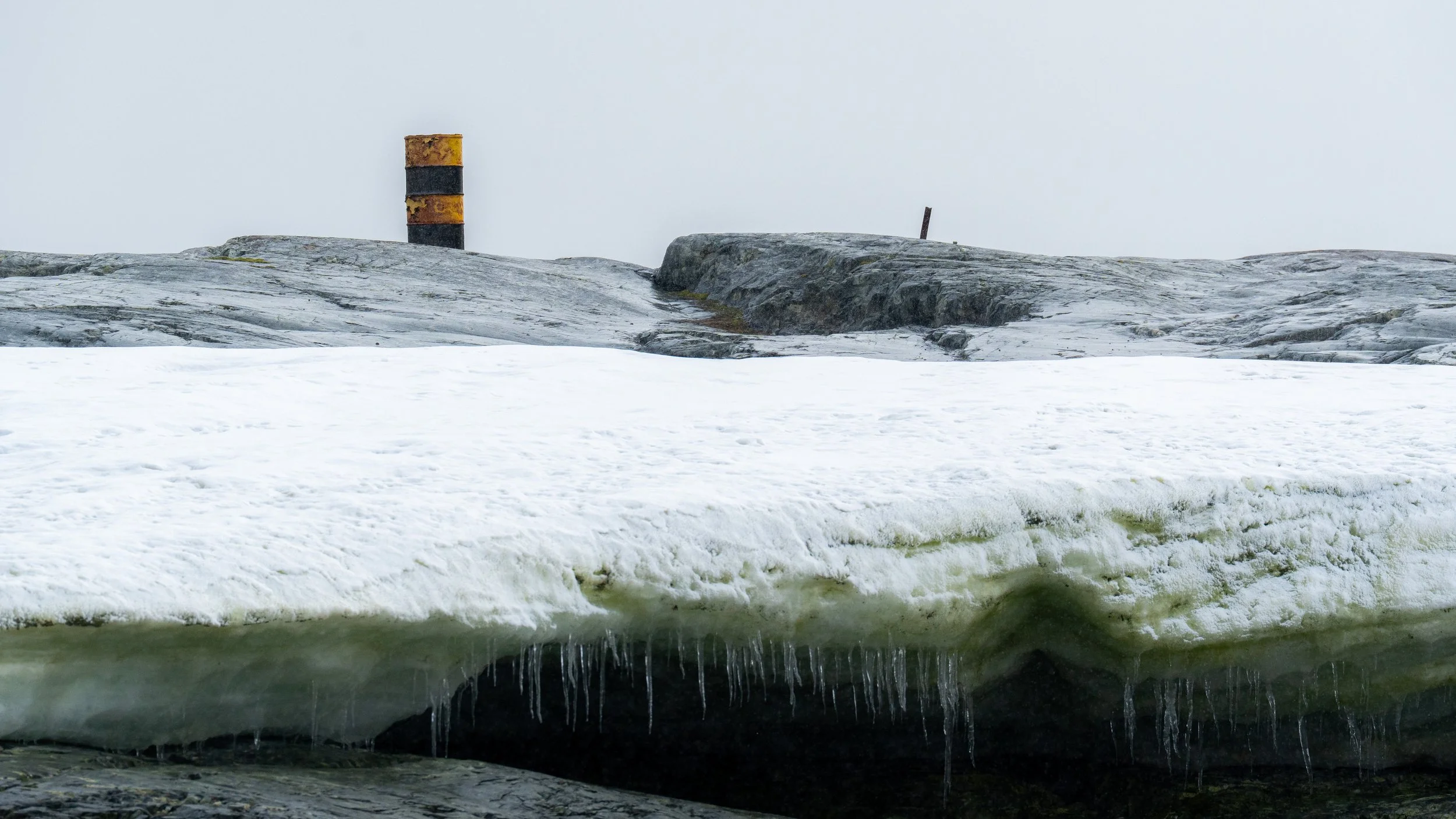 Frozen body of water with ice and snow, featuring icicles hanging underneath, and a rocky, snow-covered shoreline in the background with a yellow and black striped pole.