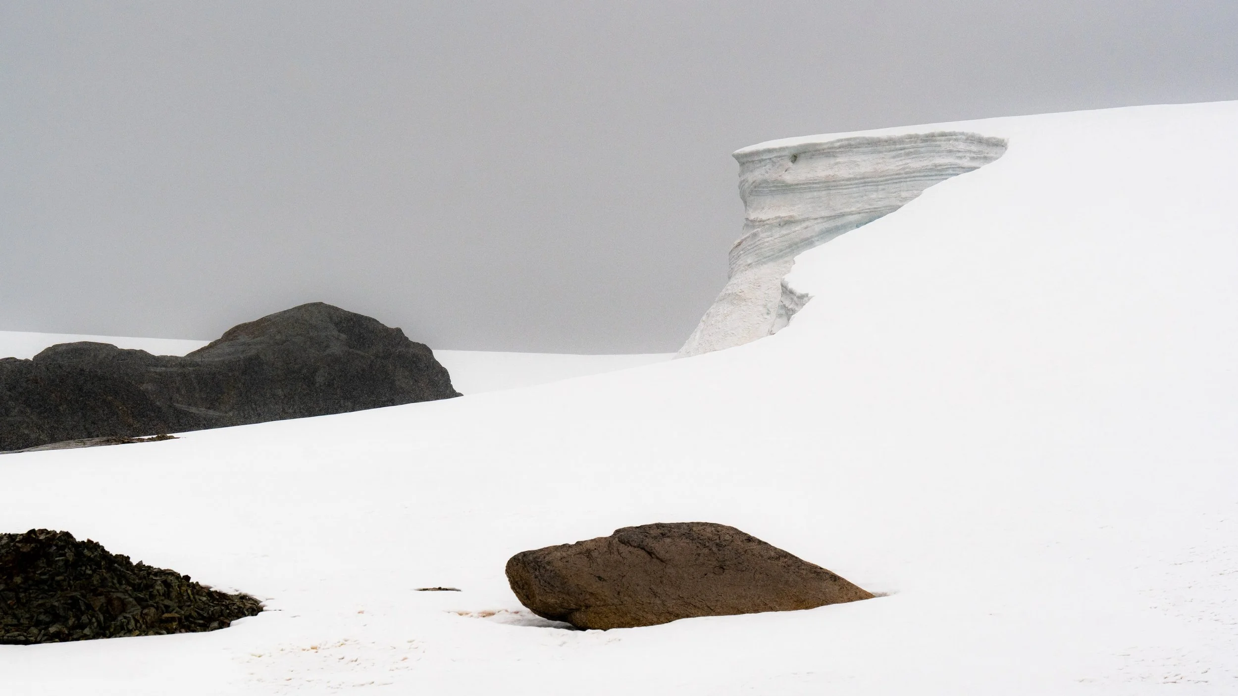 Snow-covered landscape with dark rocks in the foreground and large snow and ice formations in the background, under a gray overcast sky.