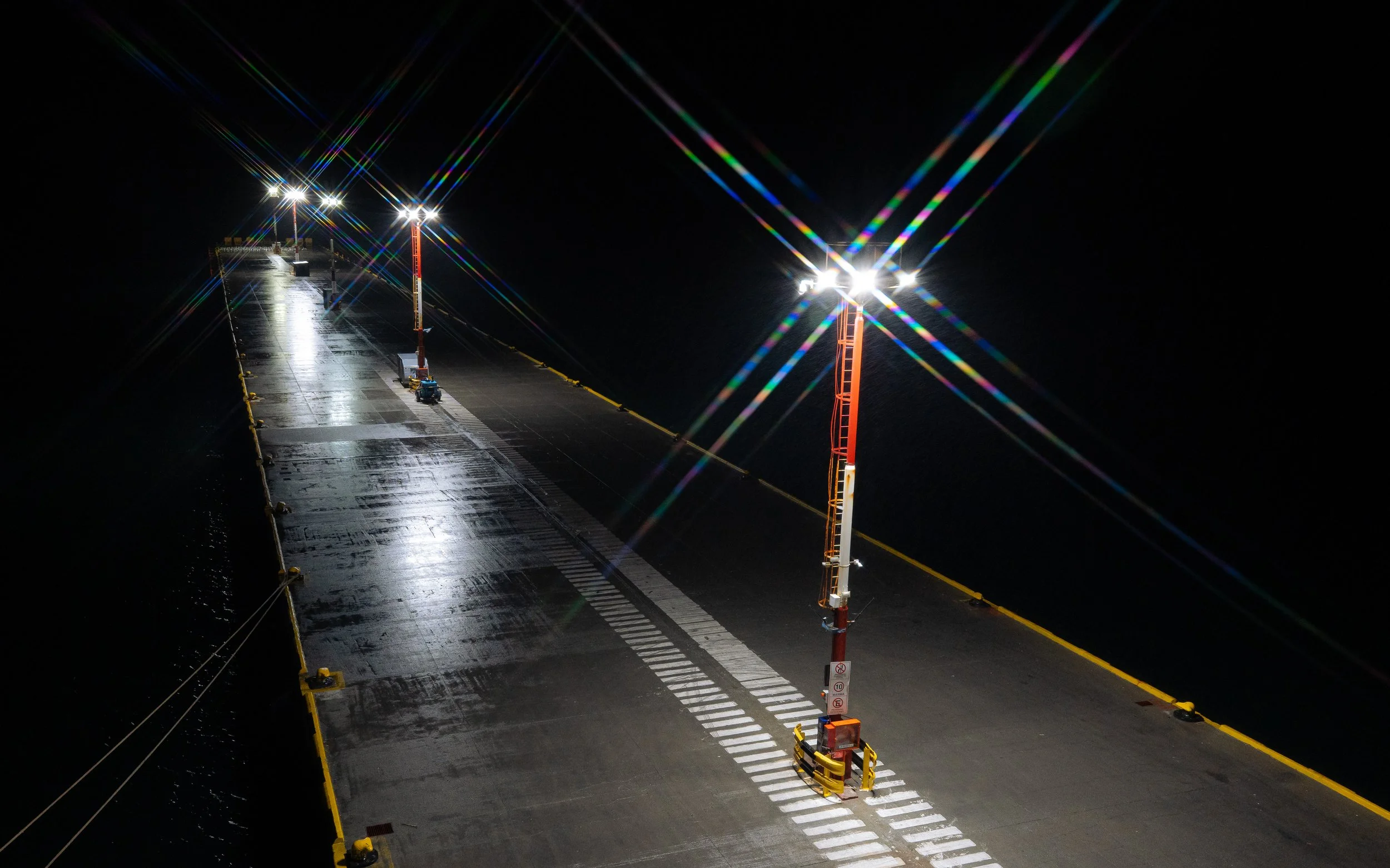 Nighttime scene of a pier with bright lights on tall orange poles, wet surface reflecting the lights, and yellow safety barriers along the edge.