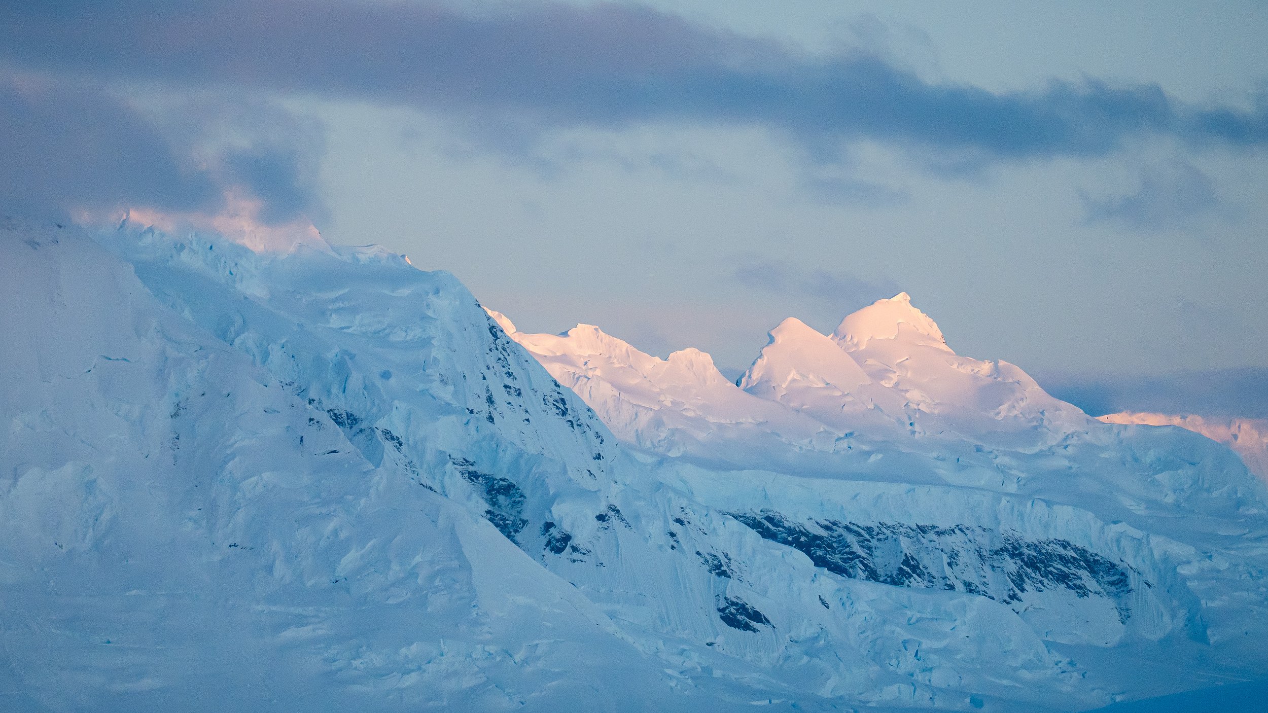 Snow-covered mountain peaks under a cloudy sky, illuminated by soft sunlight.
