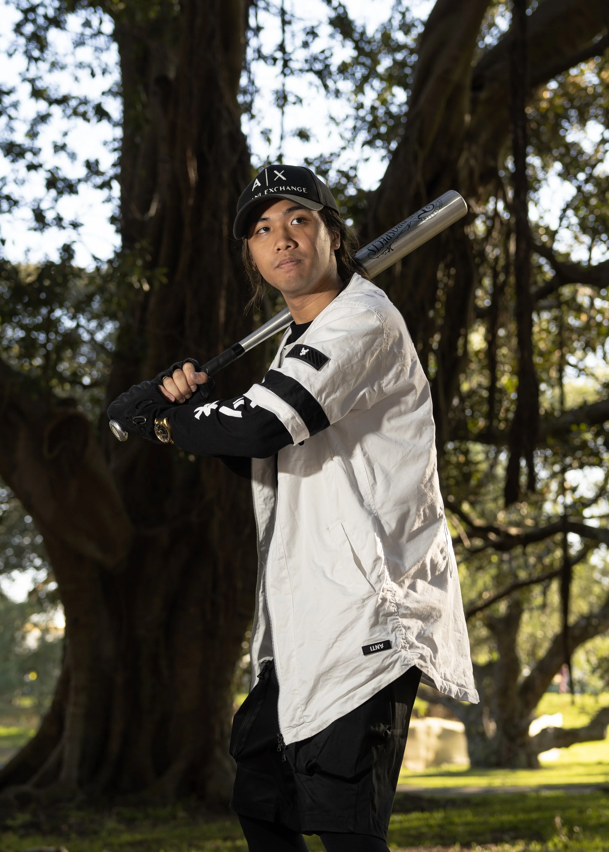 Young man holding a baseball bat over his shoulder, standing outdoors near a large tree, wearing a black cap and sports jacket.