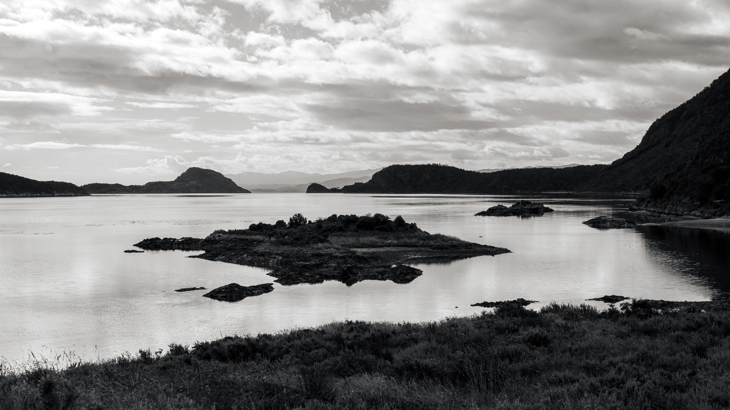 Black and white landscape of a lake with small islands and surrounding hills under a cloudy sky.