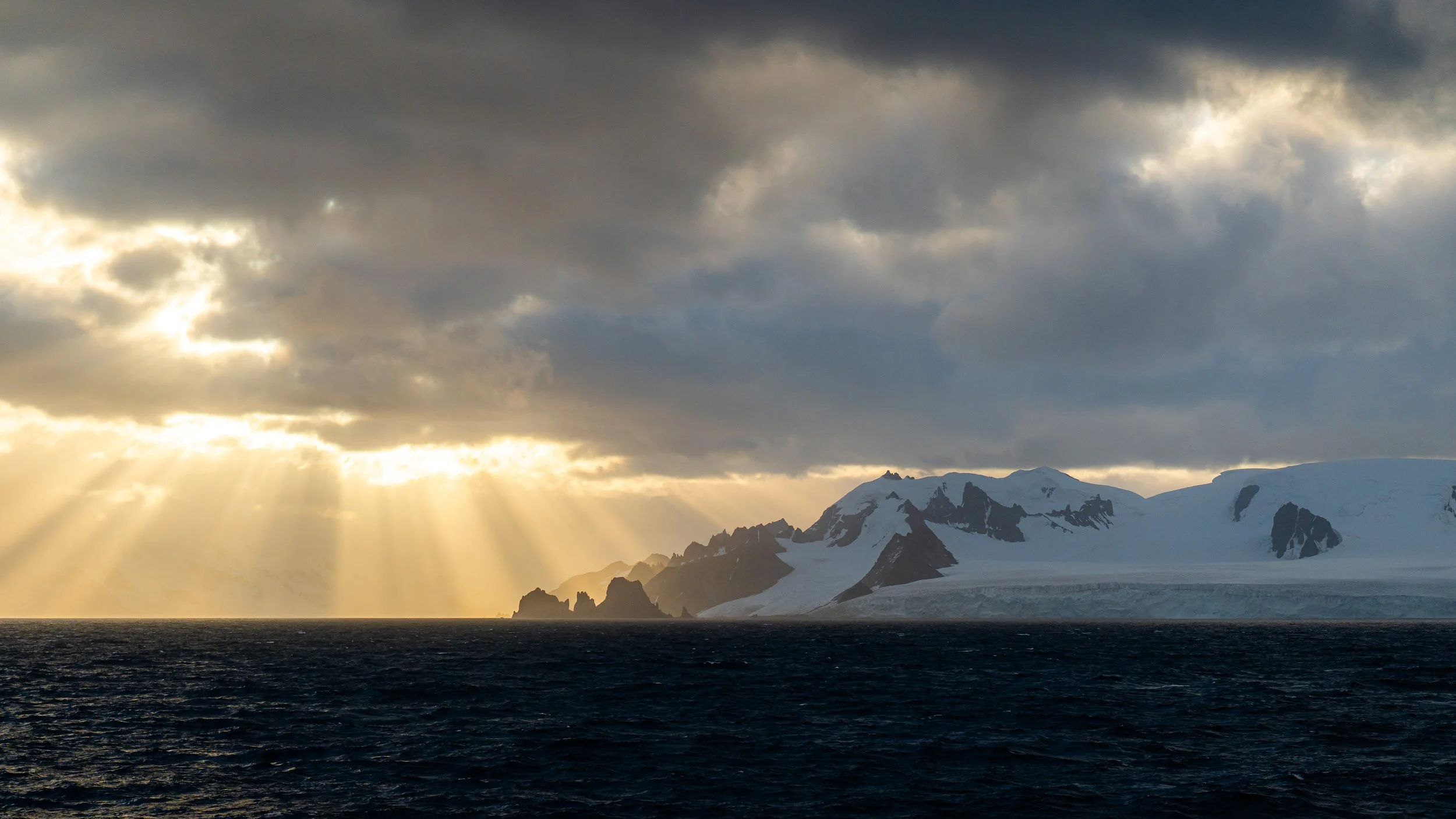 Sun rays breaking through dark clouds over snow-capped mountains and ocean.