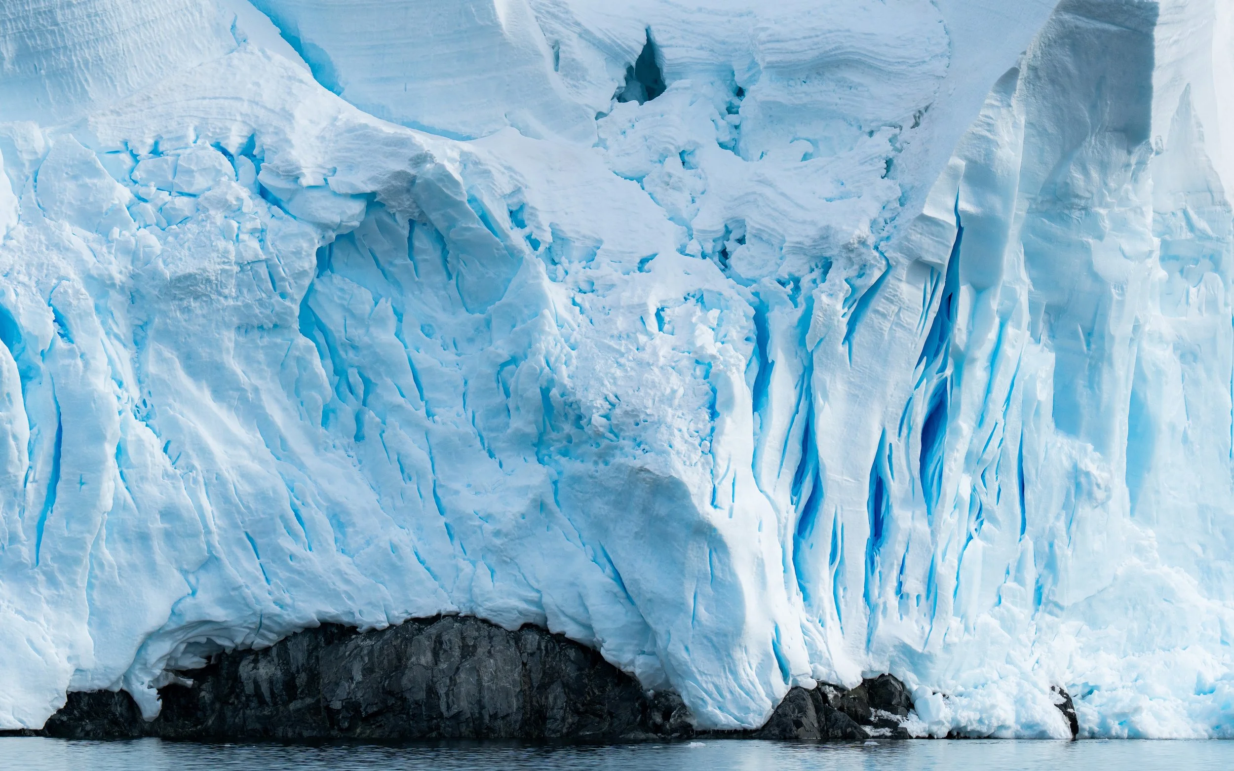Close-up of a glacier with jagged ice and crevasses, meeting dark rocky land below and reflecting in water.