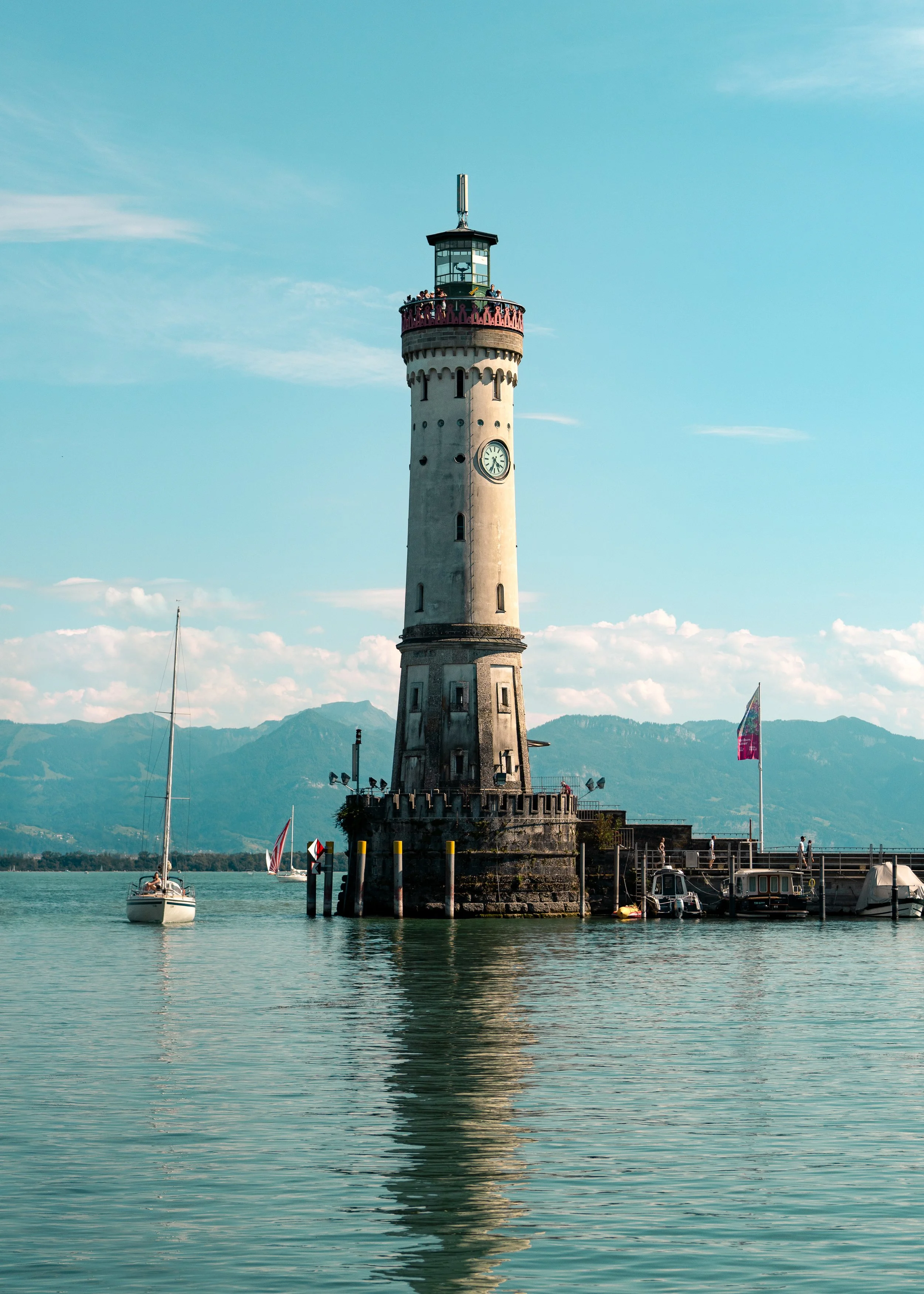 A lighthouse on the water with sailboats nearby and mountains in the background under a blue sky.