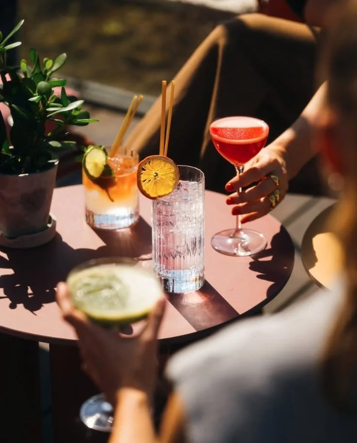 Drie mensen met cocktails op een terras met zonlicht, één persoon houdt een limoen, een andere persoon houdt een rode cocktail, en in het midden staat een glas met een citroenappelachtig drankje, met een plant op tafel.