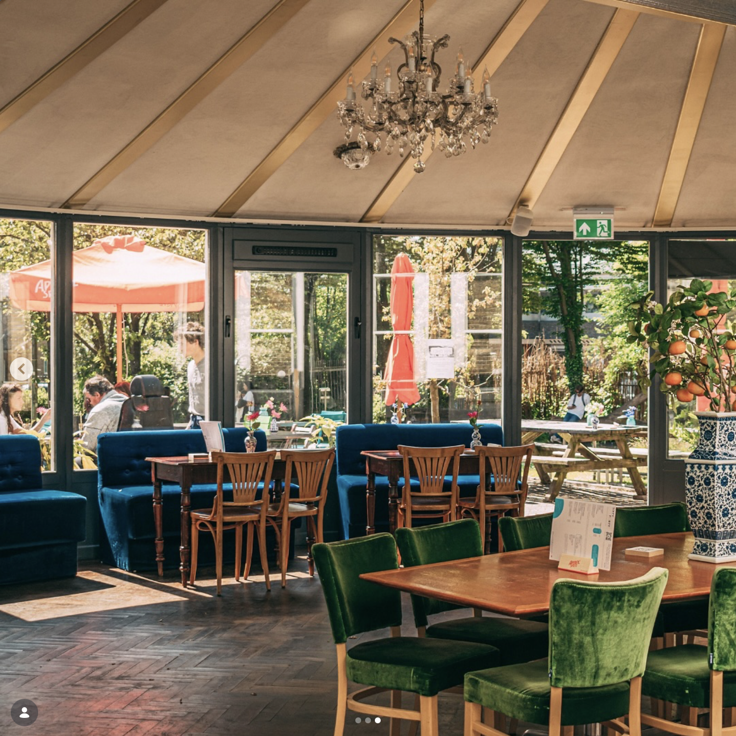 Interieur van een restaurant met groene en blauwe stoelen, houten tafels en grote ramen die uitkijken op een tuin met parasols en mensen die buiten zitten.