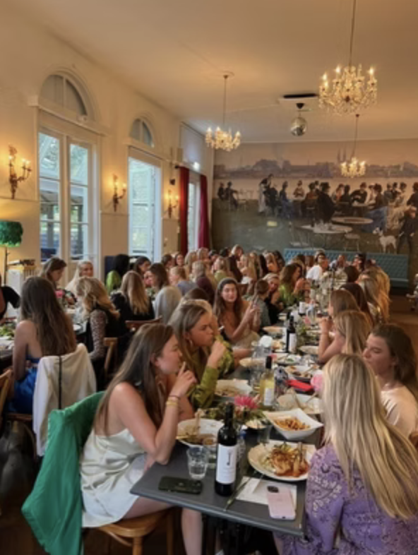 Groep jonge vrouwen zit samen aan lange tafel in restaurant met chandelier verlichting, muurkunst en grote ramen.