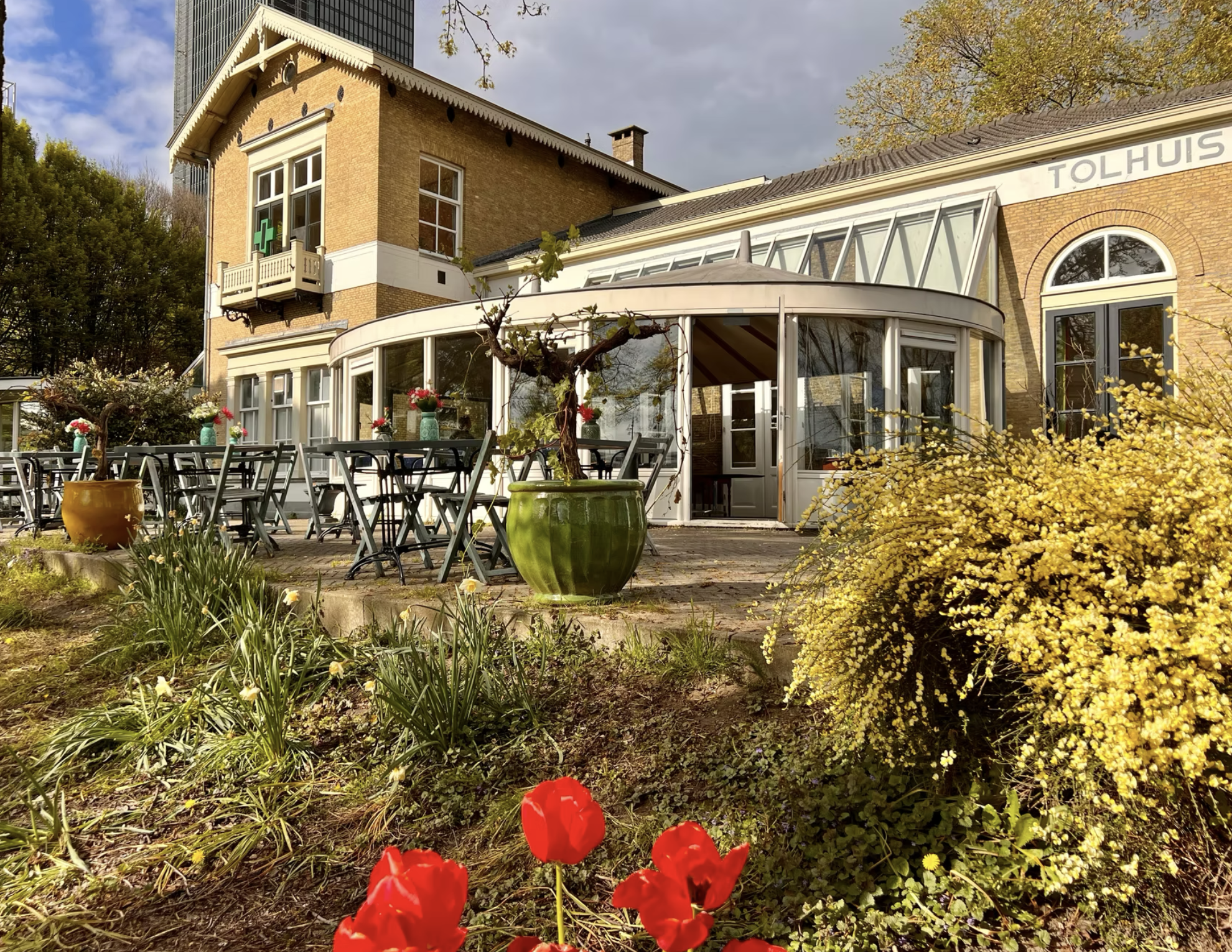 Een restaurant of terras met meerdere planten en bloemen, inclusief rode tulpen en grote potten, met een gebouw op de achtergrond dat er historisch uitziet en een kruis symbool op de gevel, mogelijk een apotheek of gezondheidscentrum, onder een bewol