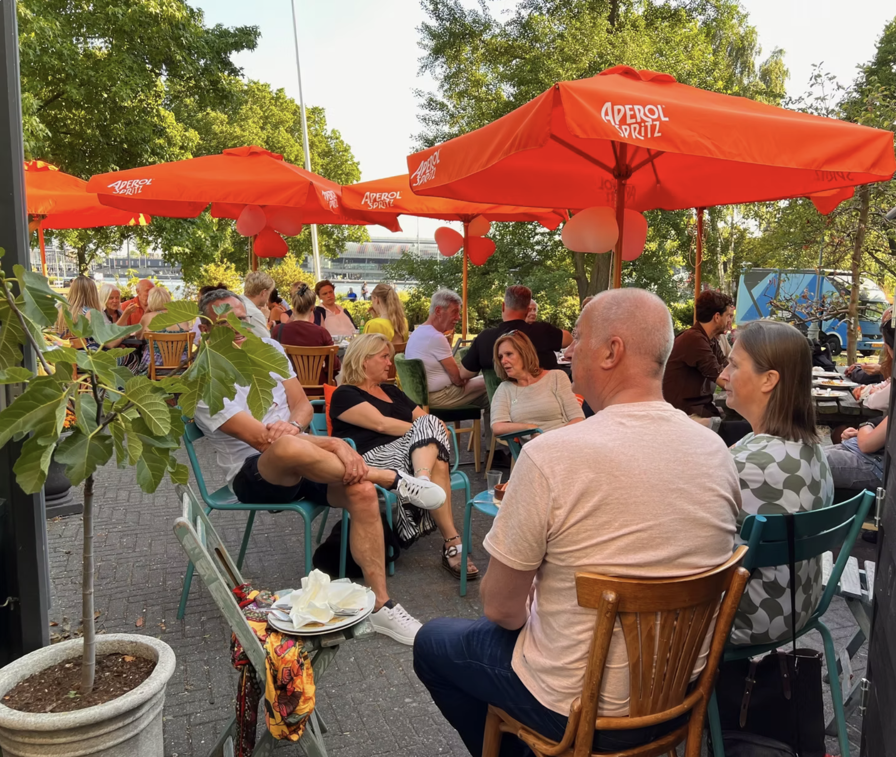 Buitenterras met mensen die praten en eten onder oranje parasols met 'Aperol Spritz' logo, groene bomen en een blauwe bus op de achtergrond.