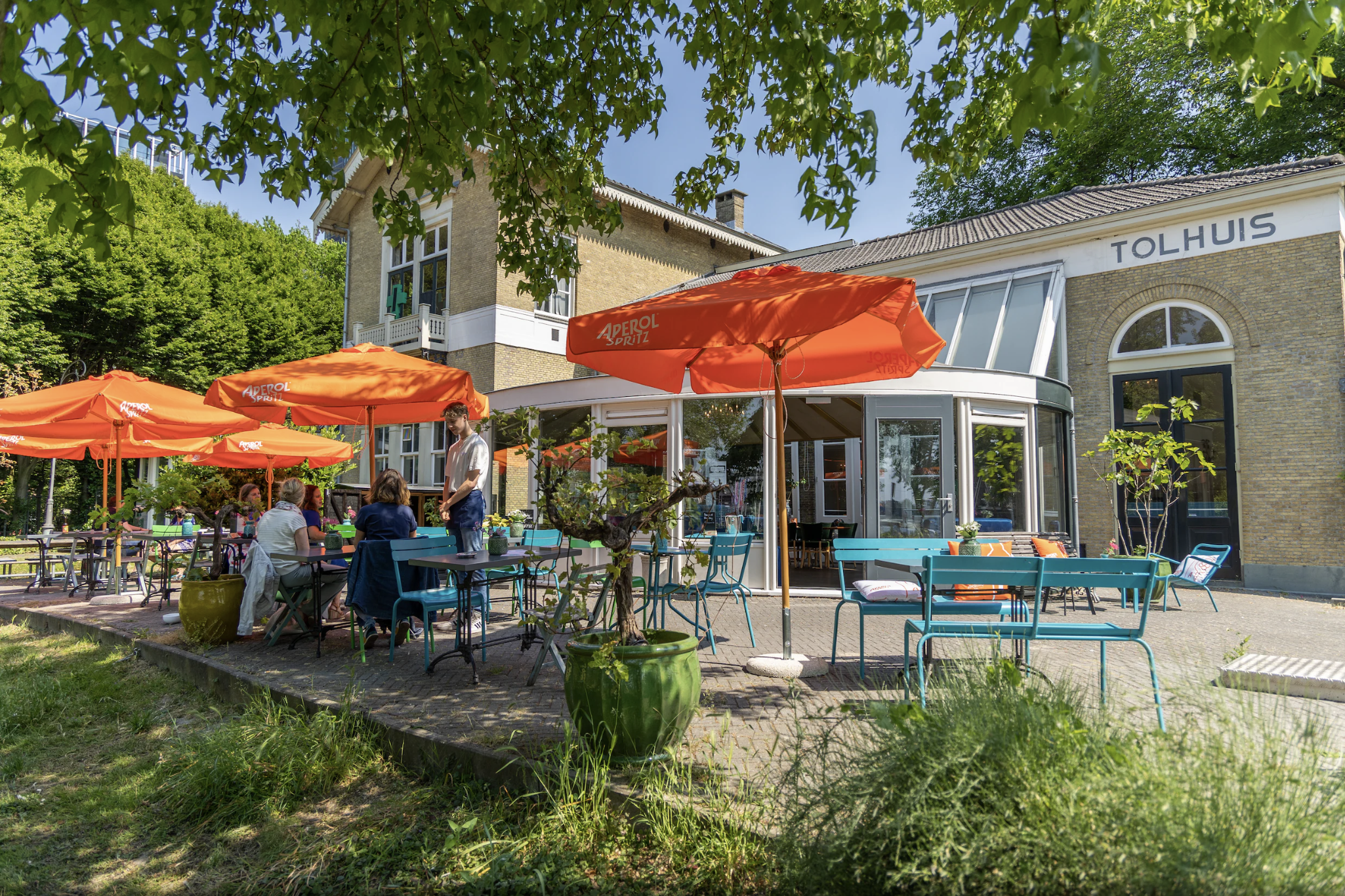 Buiten terras met kleurrijke stoelen en parasols, met mensen die genieten van het mooie weer, voor een restaurant met de naam 'Tolhuis' in een groene omgeving.
