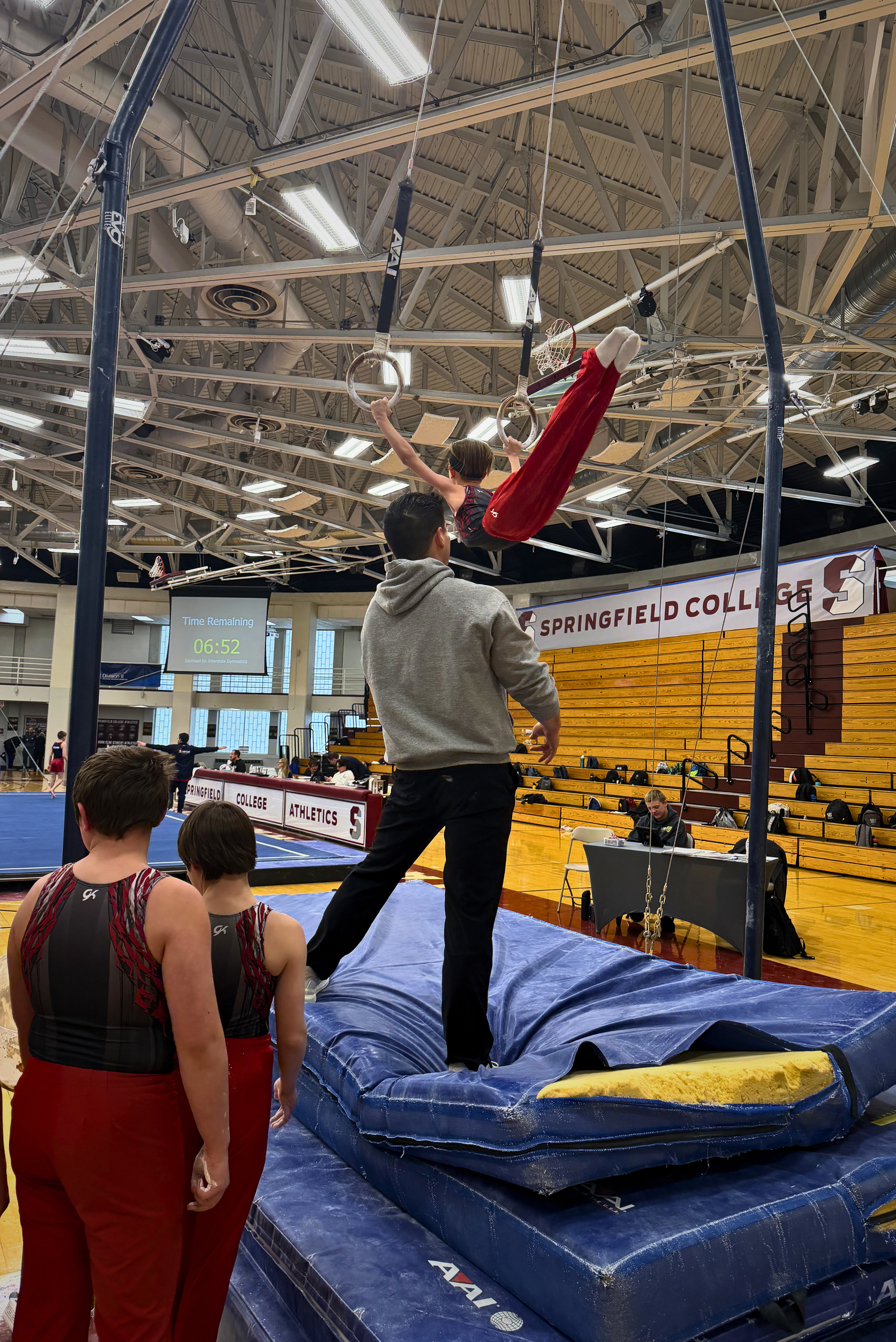 Two young gymnasts in red and black athletic attire standing on a gymnastics mat, watching a male gymnast perform on the uneven bars in a gymnasium at Springfield College. The male gymnast is in mid-air, holding gymnastic rings, wearing red pants and a sleeveless top. A digital timer on the wall shows 6 minutes and 52 seconds remaining. The gymnasium has a high ceiling with exposed beams, and a wooden bleacher section is visible. There are spectators and officials present.