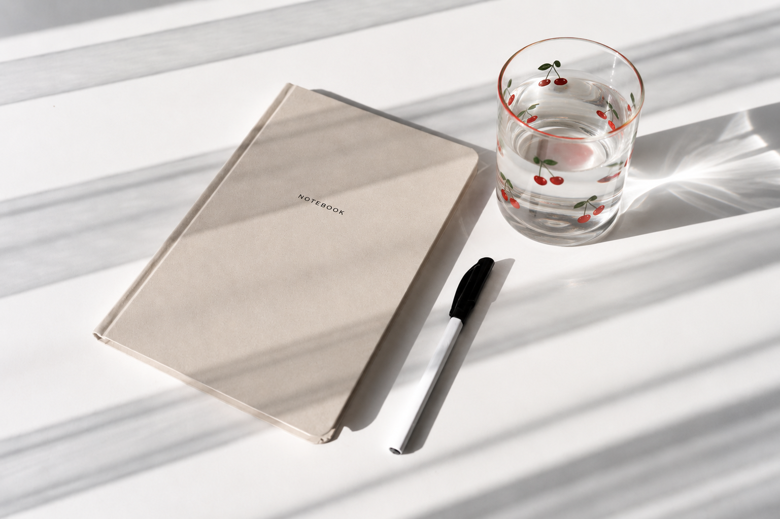 Beige notebook, black pen, and glass of water in a glass cup with a cherry design on a white tabletop in natural sunlight.