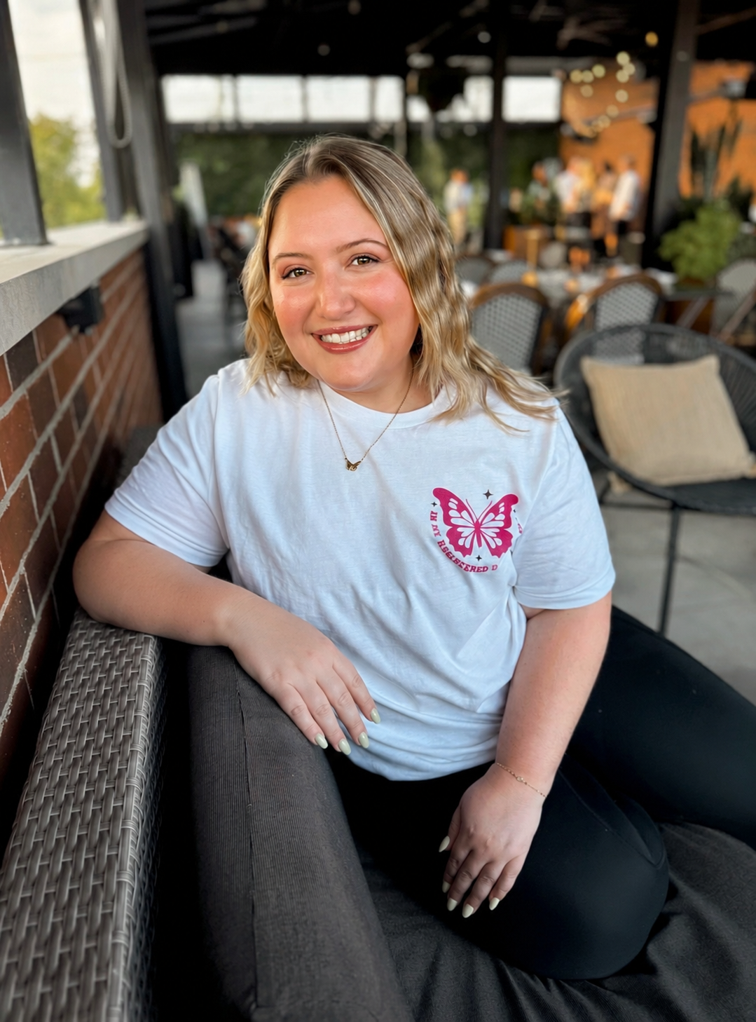 A woman sitting at an outdoor patio, smiling, wearing a white T-shirt with a pink butterfly graphic and text, with blond wavy hair and light makeup.