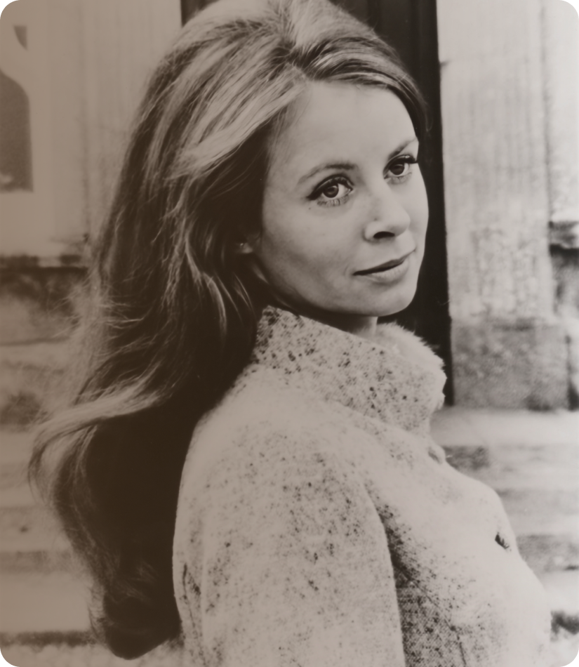 Black and white photo of a woman with long wavy hair, wearing a knitted sweater, sitting outdoors near a wooden porch, looking over her shoulder at the camera.