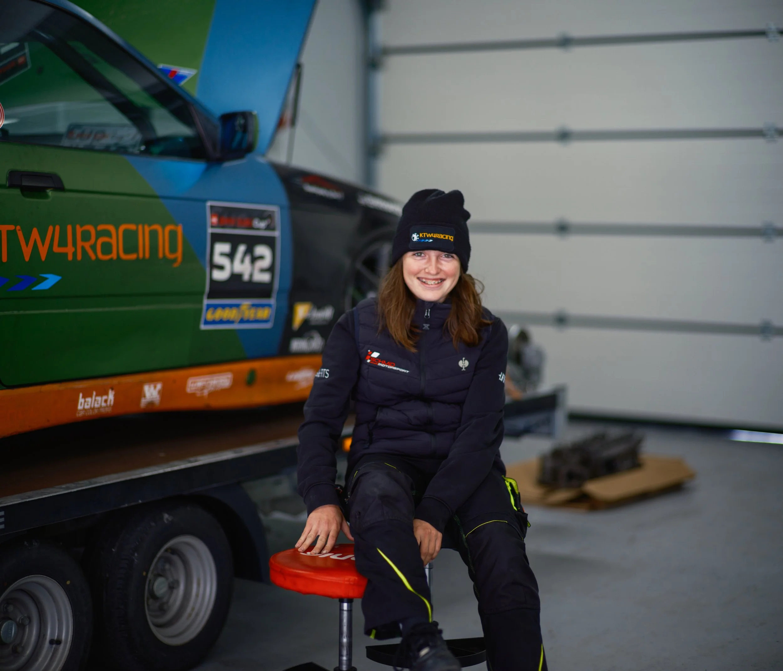 A woman with long red hair smiling while sitting in a racing car trailer, wearing a black beanie and black racing suit, with her hand on a red racing helmet, in a garage or workshop.