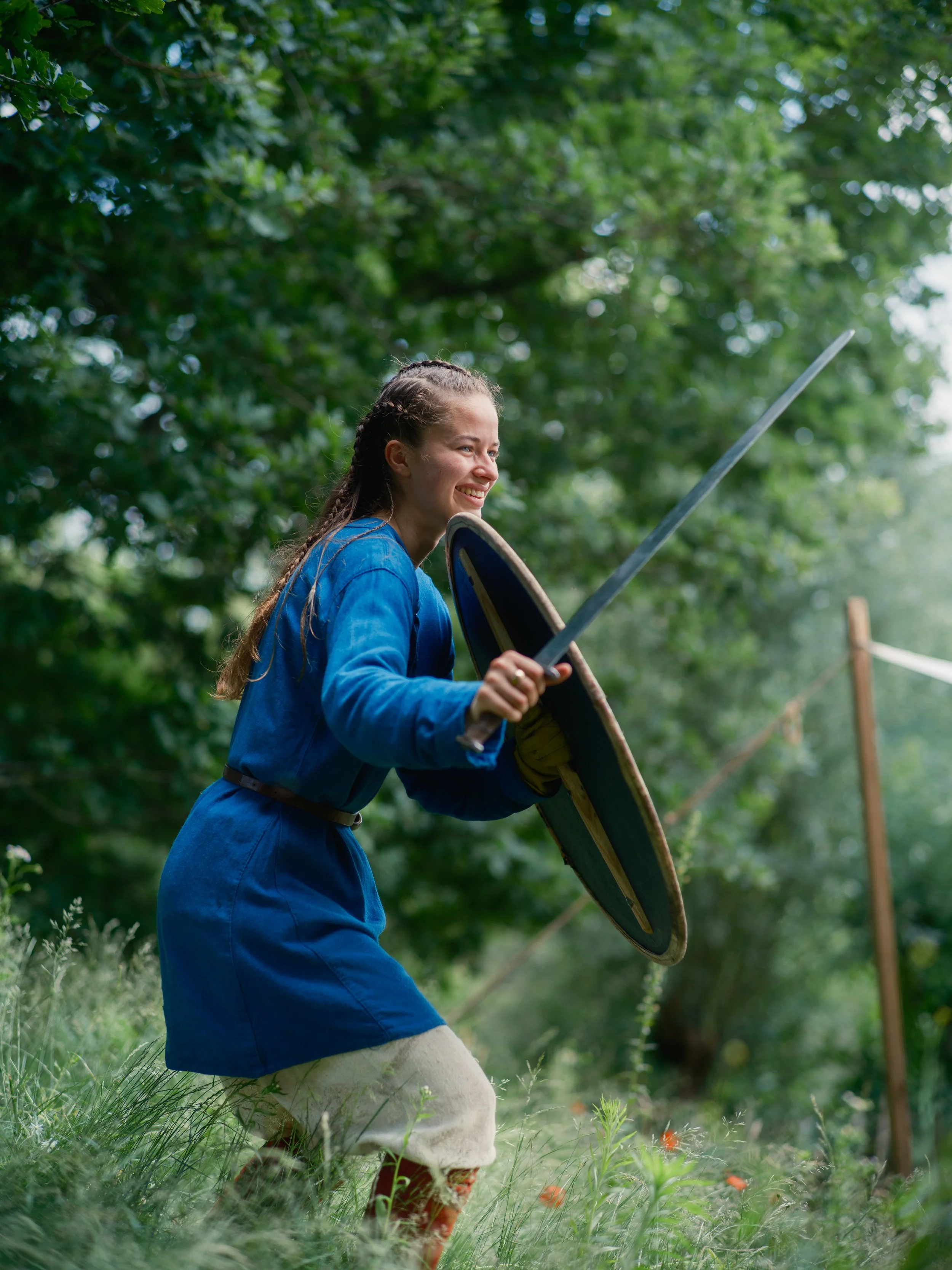 A young woman dressed in medieval or fantasy-style clothing, kneeling in a lush green outdoor forest setting, holding a round shield and a long sword, smiling.