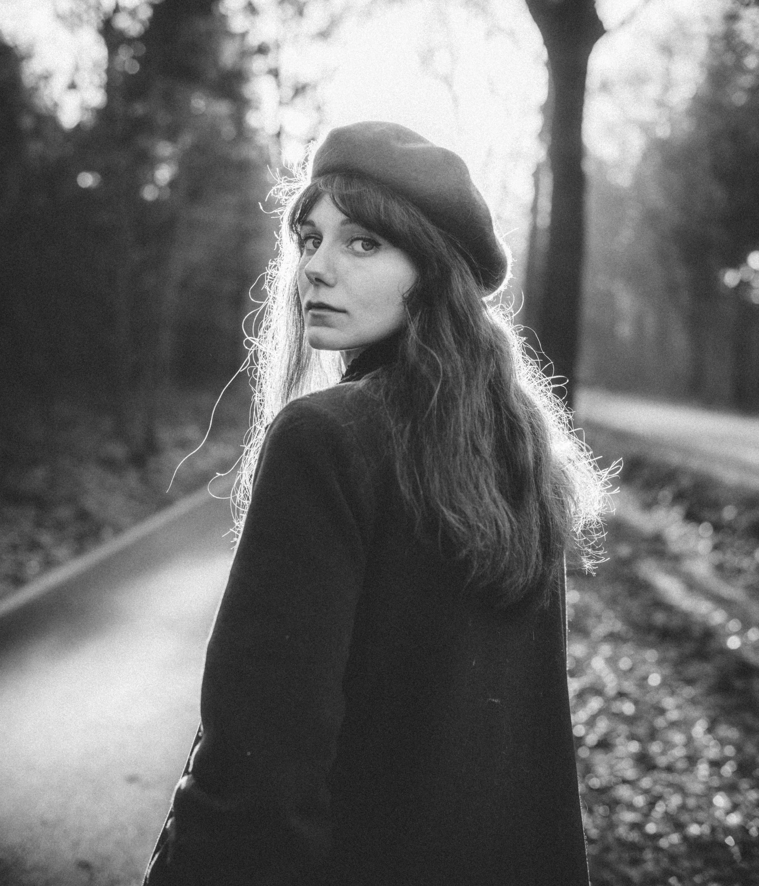 A black and white photo of a woman with long hair, wearing a beret, standing outdoors with a blurred background of trees and light filtering through.