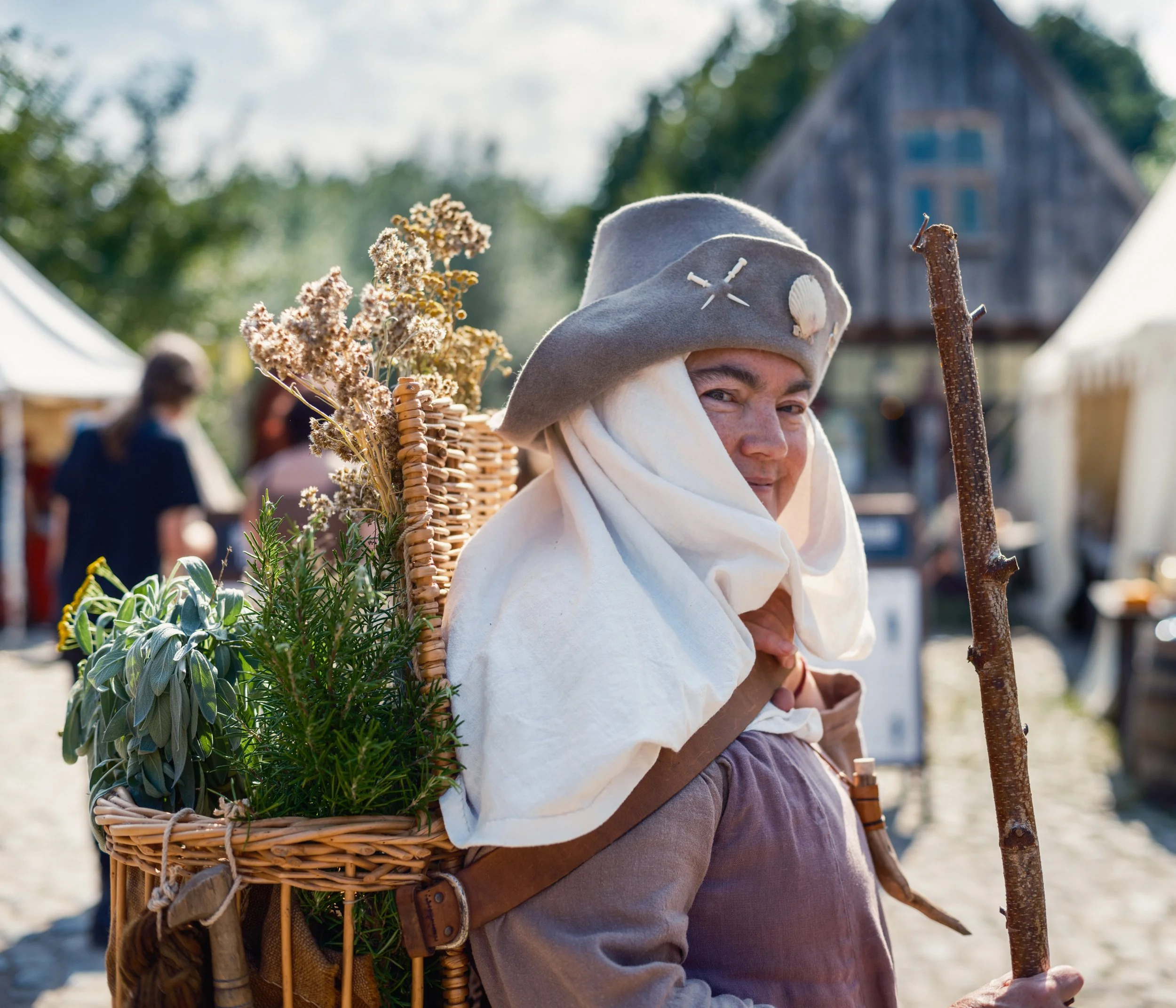 A person dressed in medieval costume smiling while holding a staff, wearing a grey hat with small shells and stitching, carrying a wicker basket filled with dried flowers and herbs, outdoors with blurred background of trees and a rustic building.