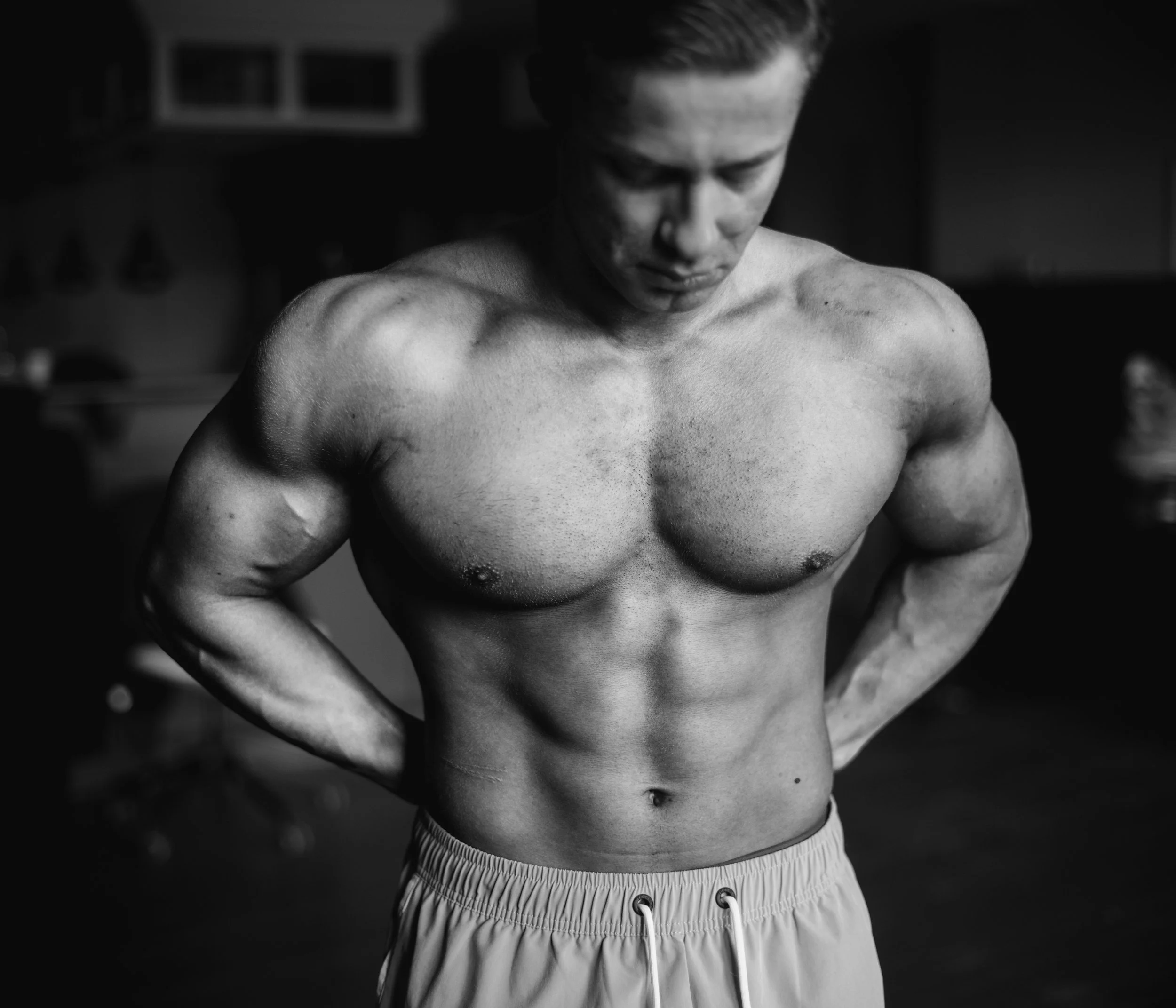 Black and white photo of a shirtless man wearing striped athletic shorts with a drawstring, standing indoors with his head tilted down.