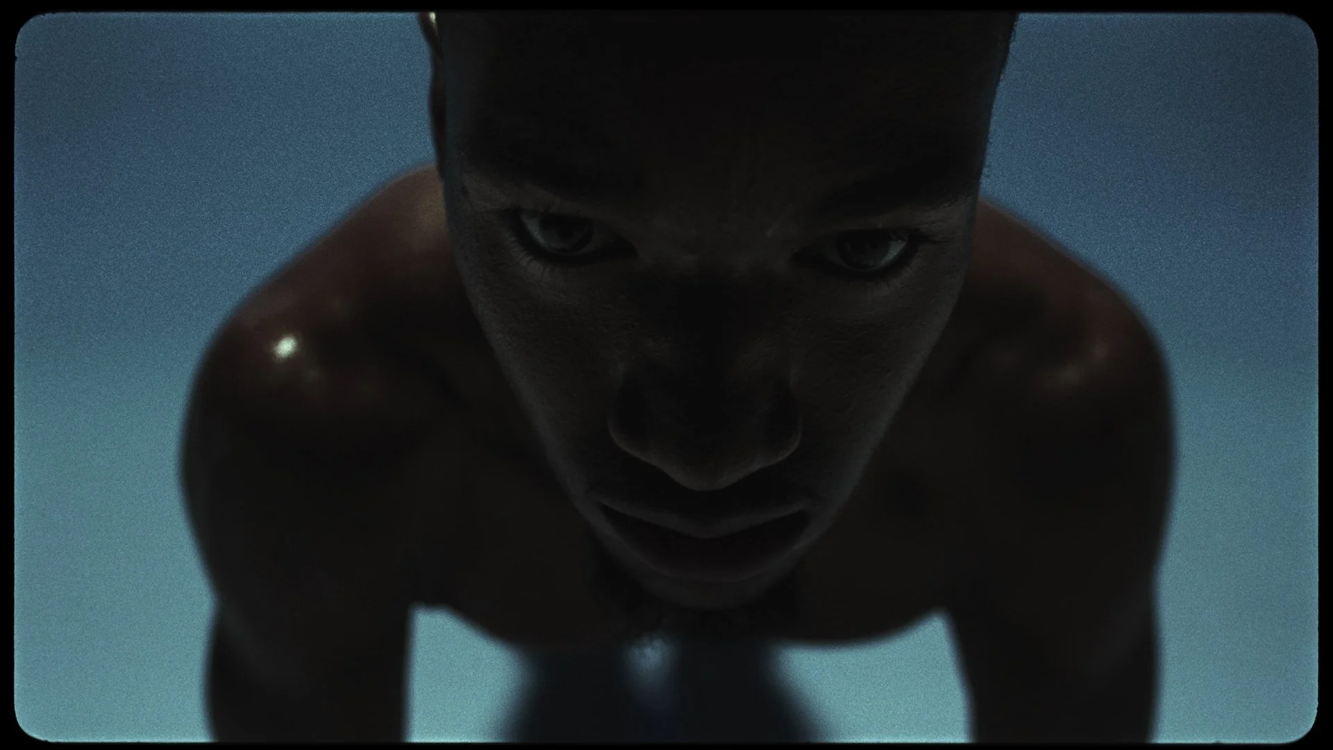 A close-up shot of a shirtless man with an intense expression, leaning forward with a blue background.