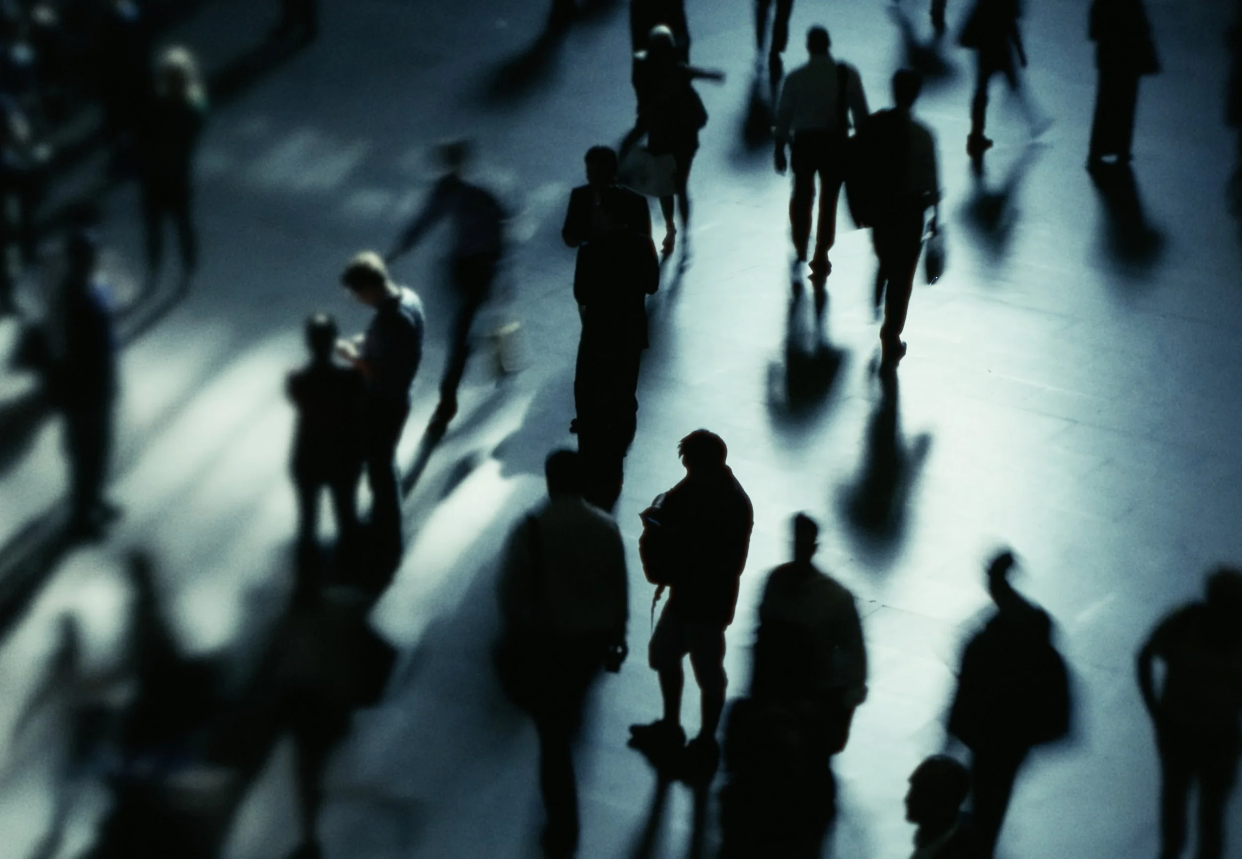 Silhouetted people walking on a glossy floor, with reflections and blurred motion, in a dimly lit environment.