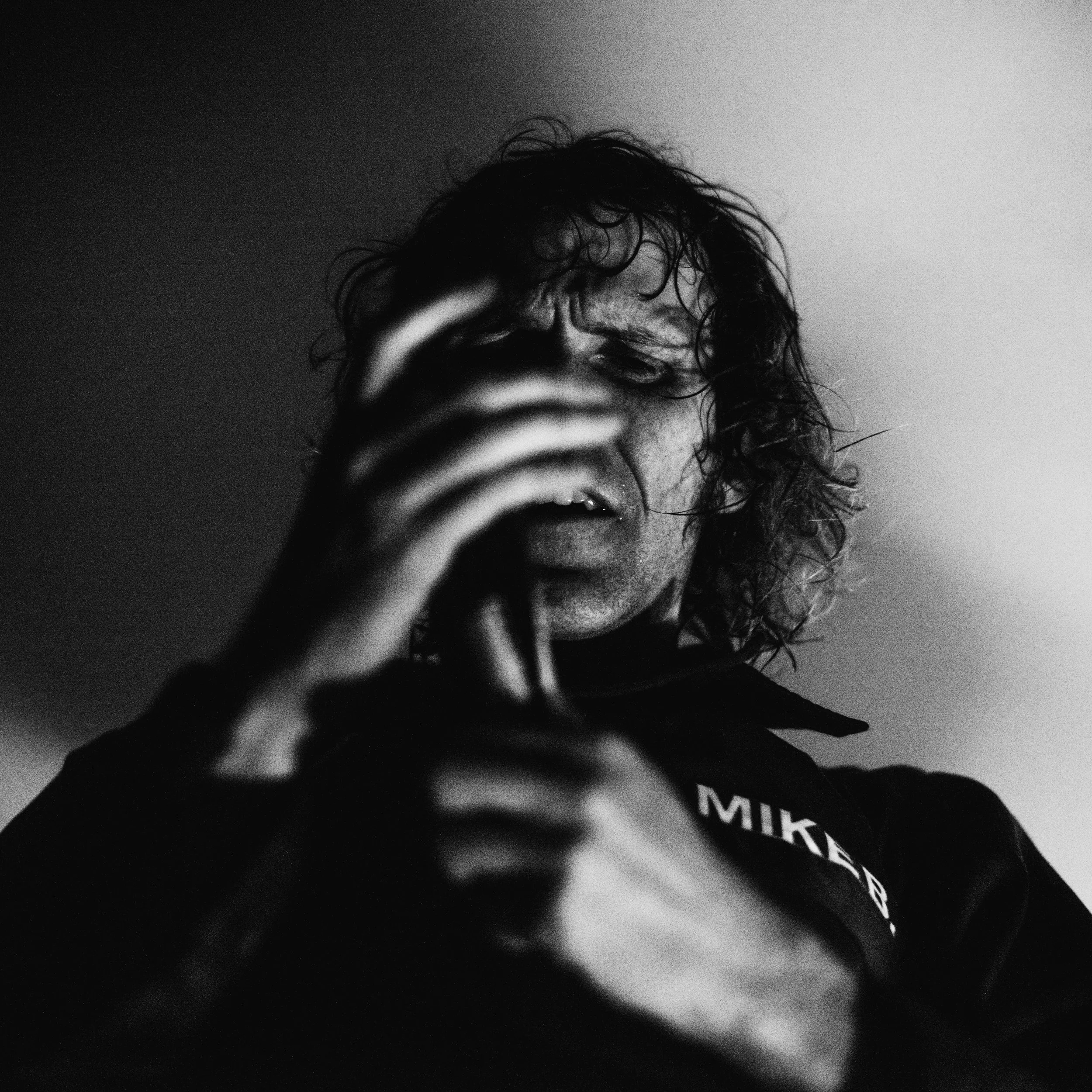 A black and white photo of a person with curly hair, holding a camera or phone in front of their face, partially covering their nose and mouth. The person's eyes are closed, and they are wearing a dark shirt or jacket.