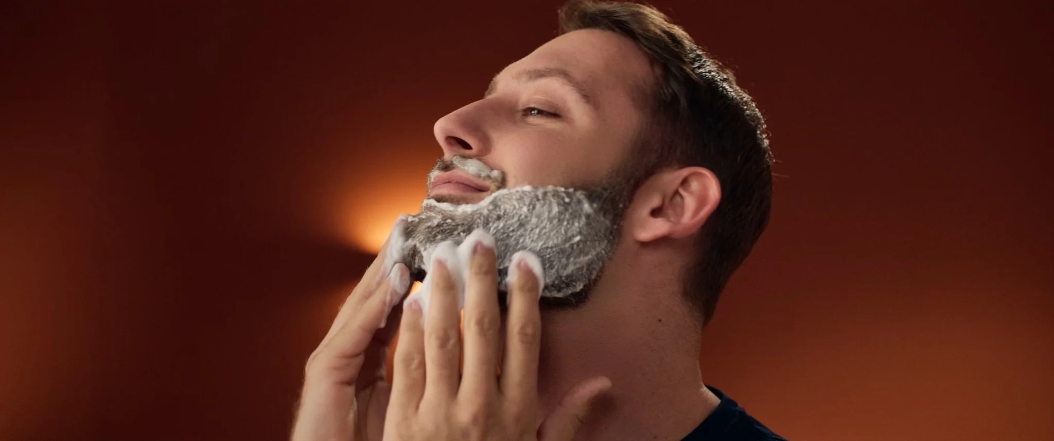 A man with light skin and short brown hair shaves his face, covered with white shaving cream, against a brown background.