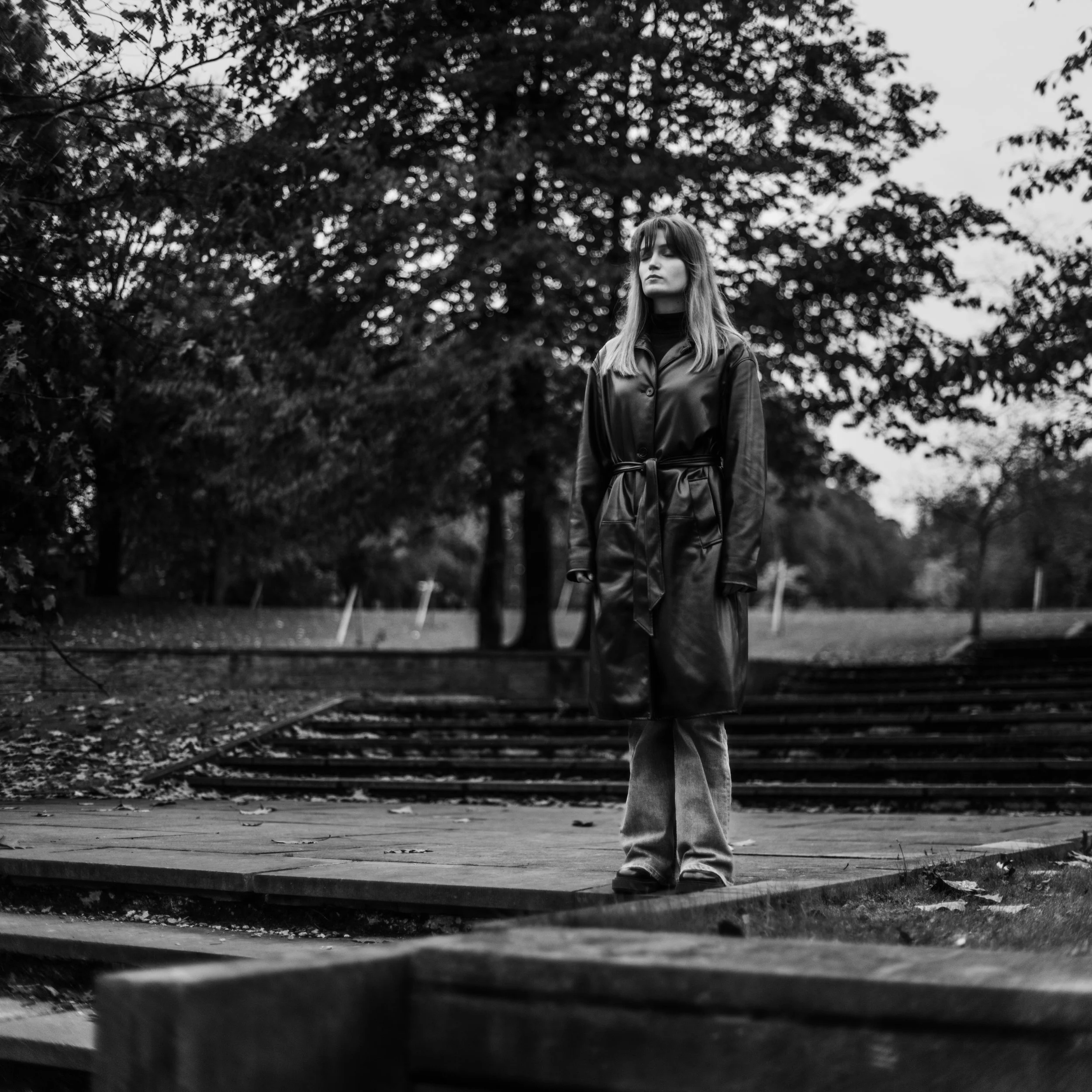 Black and white photo of a woman standing outdoors near trees, wearing a leather jacket and pants, with her eyes closed and head tilted slightly upward.