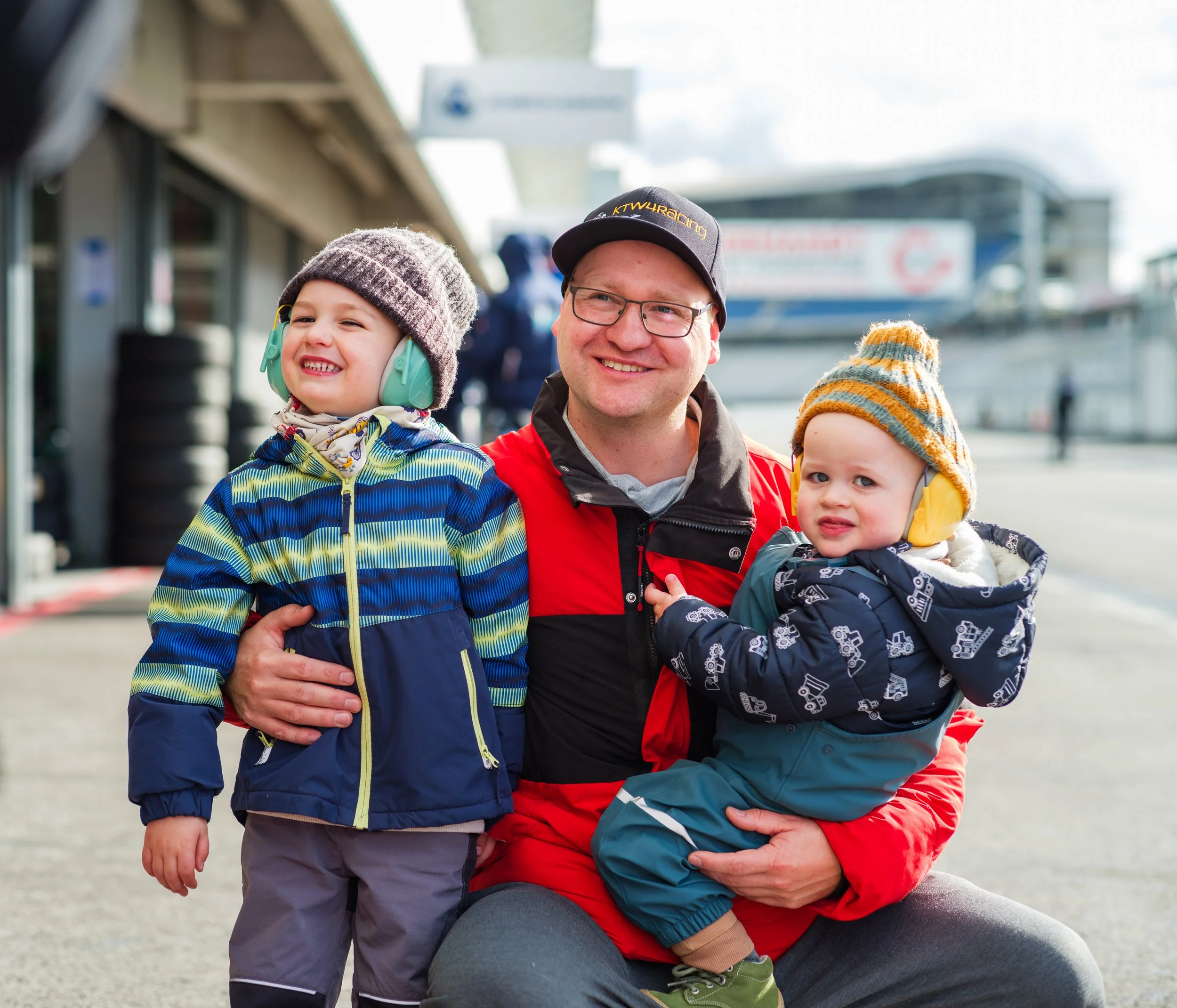 A man with glasses and wearing a hat, smiling, kneeling with two young children outdoors. The children are wearing hearing protection and colorful jackets, and are smiling or looking curious.