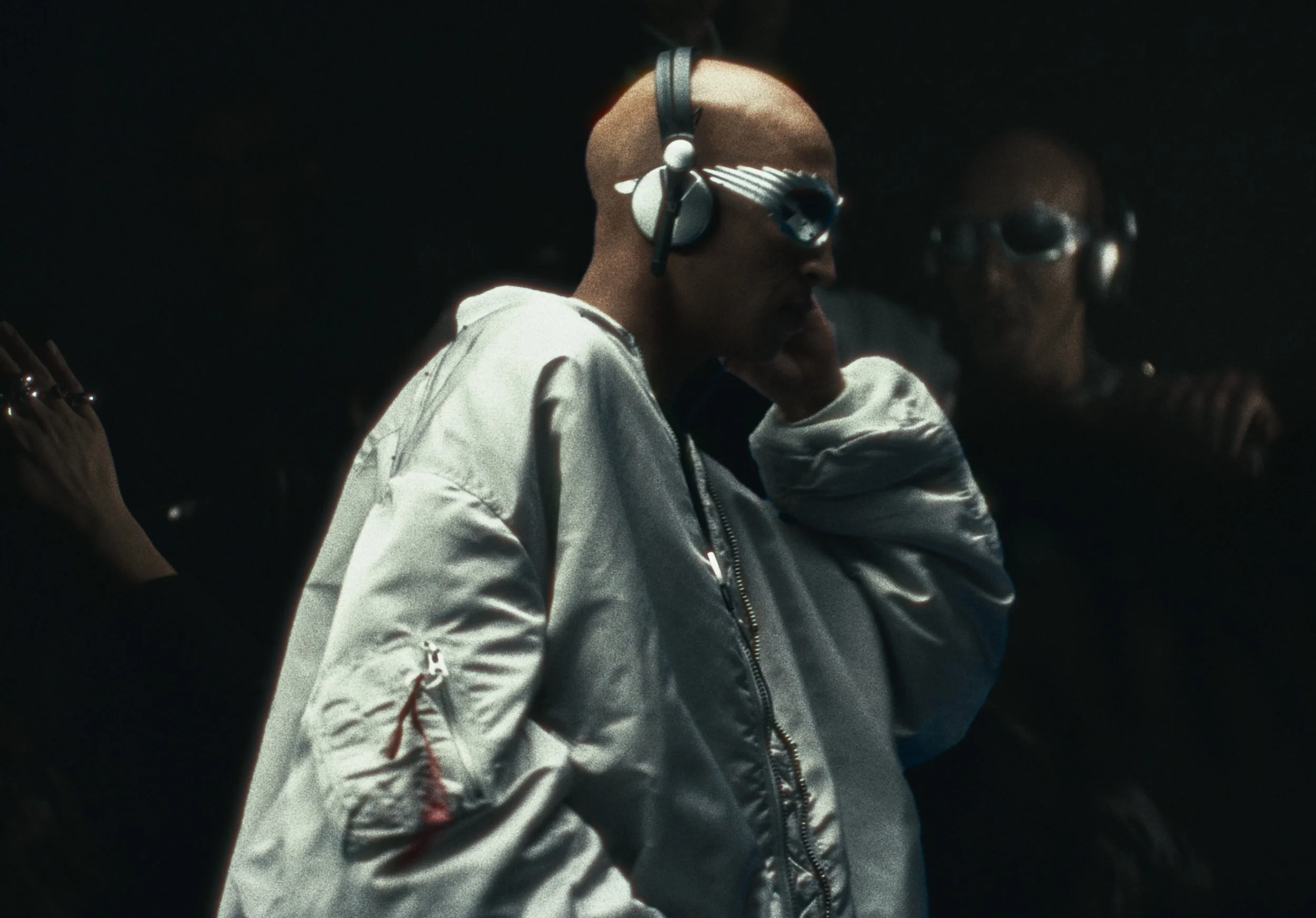 A man with a shaved head wearing large headphones and a shiny silver jacket. He is seated and appears focused, with another person partially visible next to him in a dark setting.