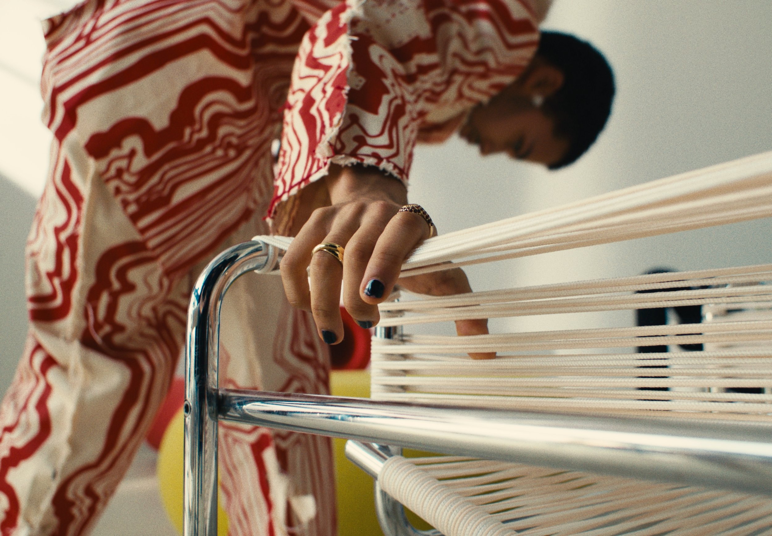 A woman with short dark hair, wearing a red and white patterned shirt and rings, is seen holding a white cloth and pulling or adjusting fabric on a clothing rack.