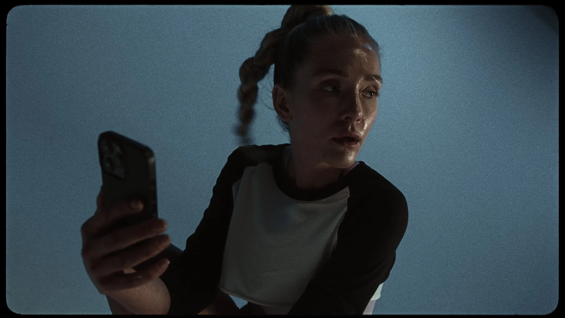 A woman with braided hair and a black and white raglan shirt is looking at her phone with a surprised or intense expression against a plain blue background.